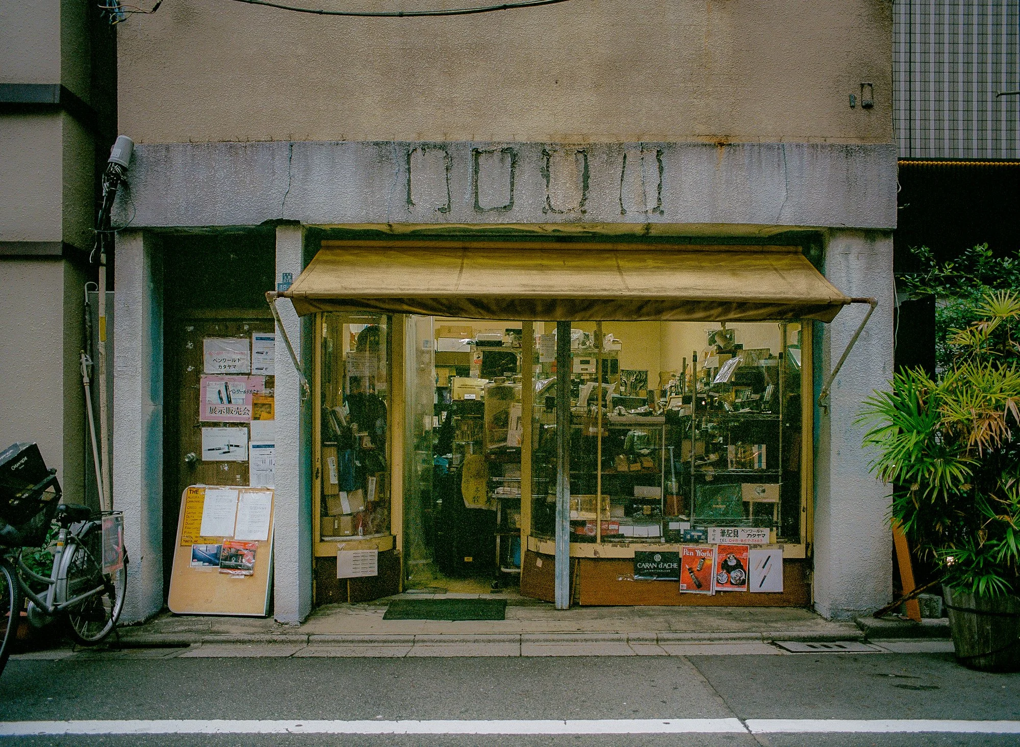 A small shop with a yellow awning, glass windows, and a wooden door, displaying high-end fountain pens inside. There is a bicycle parked on the left side of the store, and a large potted plant on the right.
