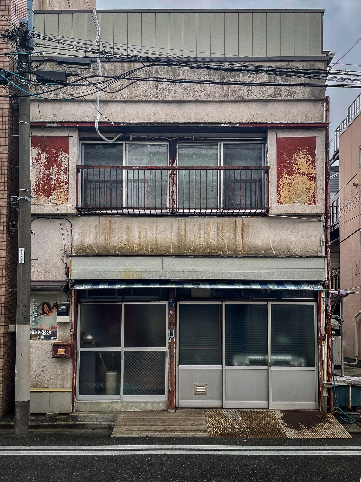 An old, weathered three-story building with a rusty balcony on the second floor, peeling paint, and a closed storefront on the ground level with glass doors and windows. The street in front has white lane markings, and electrical wires are visible ov