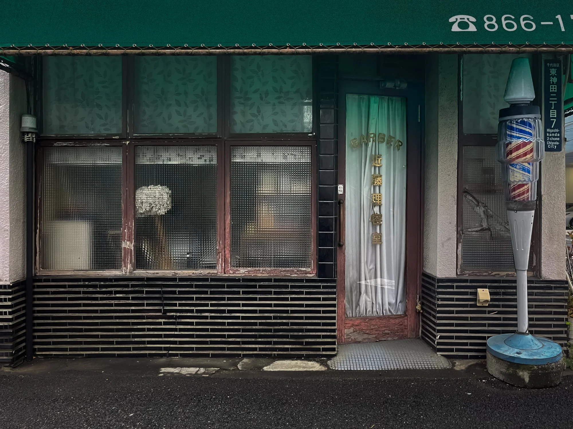Front of a small barbershop with large windows, a door with a curtain, and a vintage barber pole outside.