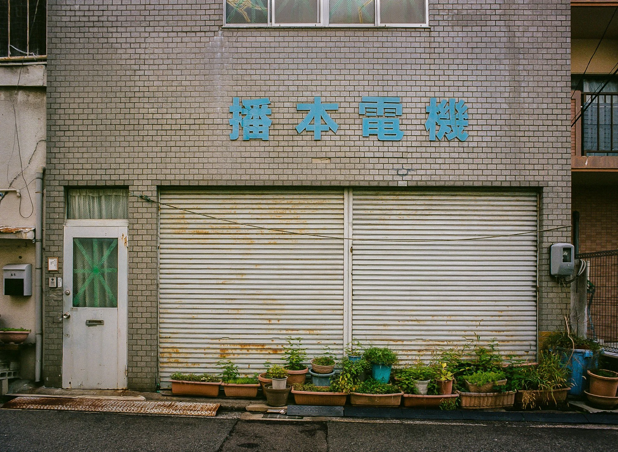A building with a brick facade and a metal rolling door, with potted plants along the bottom and Japanese characters on the upper part of the wall.