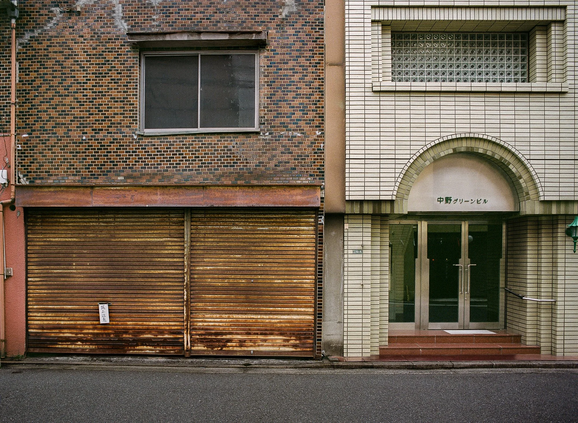 Street view of two buildings with different architectural styles, one with a brick facade and a metal shutter, and the other with white tiles and glass doors, with a small sign above the entrance.