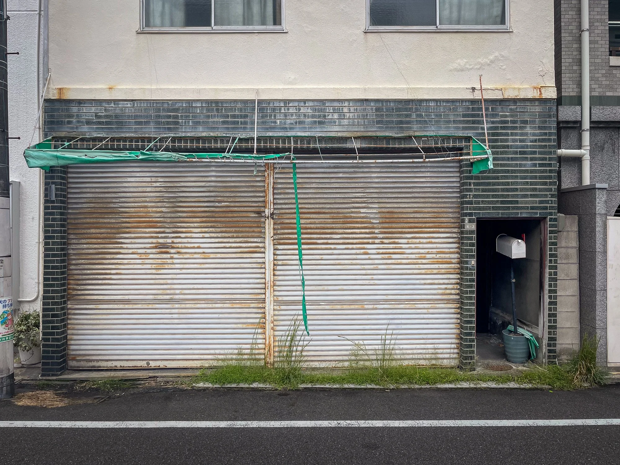 A closed, rusty metal garage door with a green cover above, set in a brick wall with a small door to the right containing a mailbox and a broom inside. The sidewalk has grass growing at the base of the garage. The street in front has white lane marki
