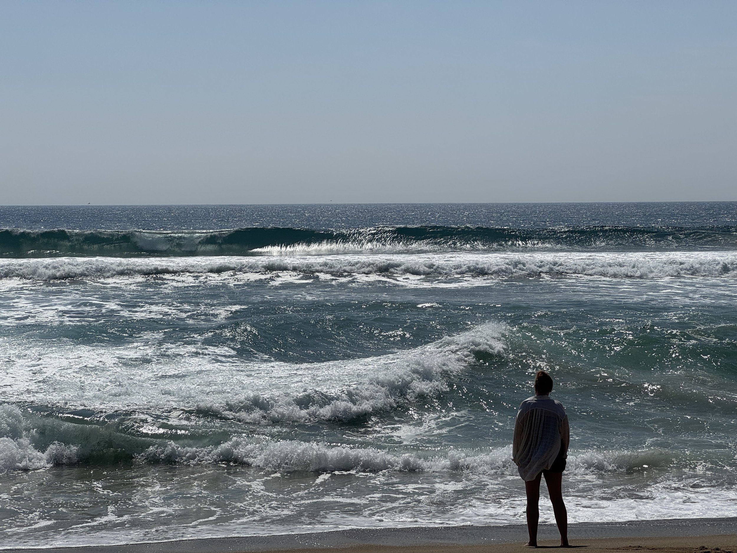 Nora Bateson stands on the sandy beach, gazing at the ocean waves under a clear blue sky.