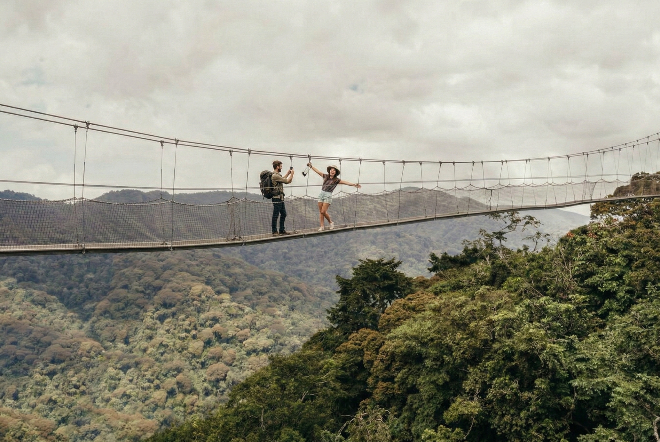 A man with a backpack photographs a woman in a hat posing with arms outstretched on a long, narrow suspension bridge above the dense tropical forest of Nyungwe National Park, Rwanda.