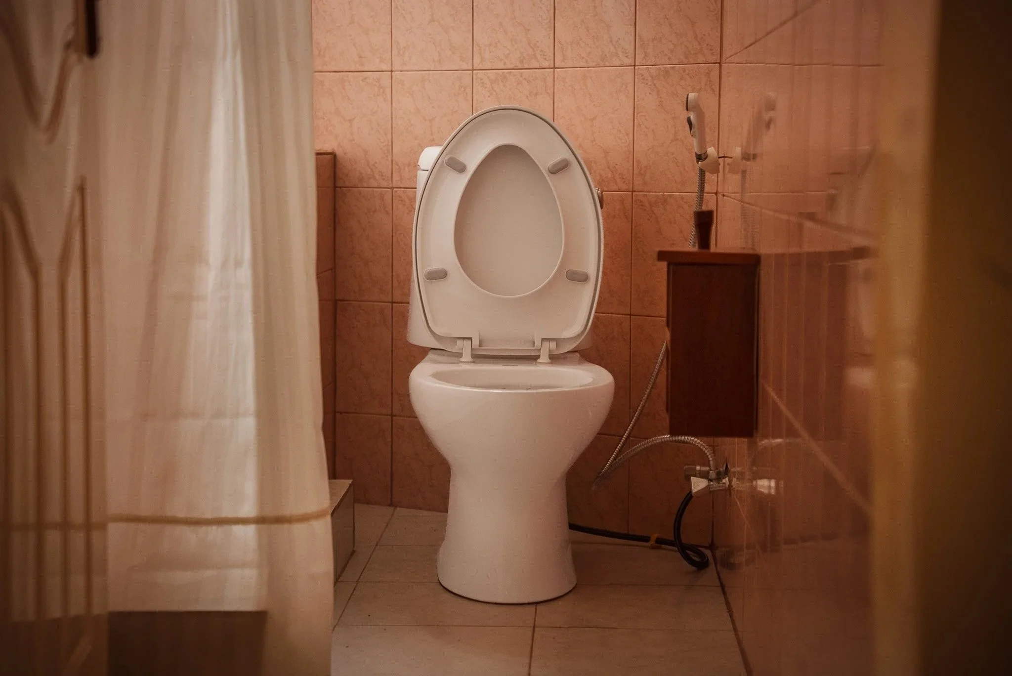 A white toilet with the lid up in a small, tiled bathroom, with a beige curtain partially visible on the left and a wooden shelf or cabinet on the right at Quality Inn Hotel Kigali, Rwanda.