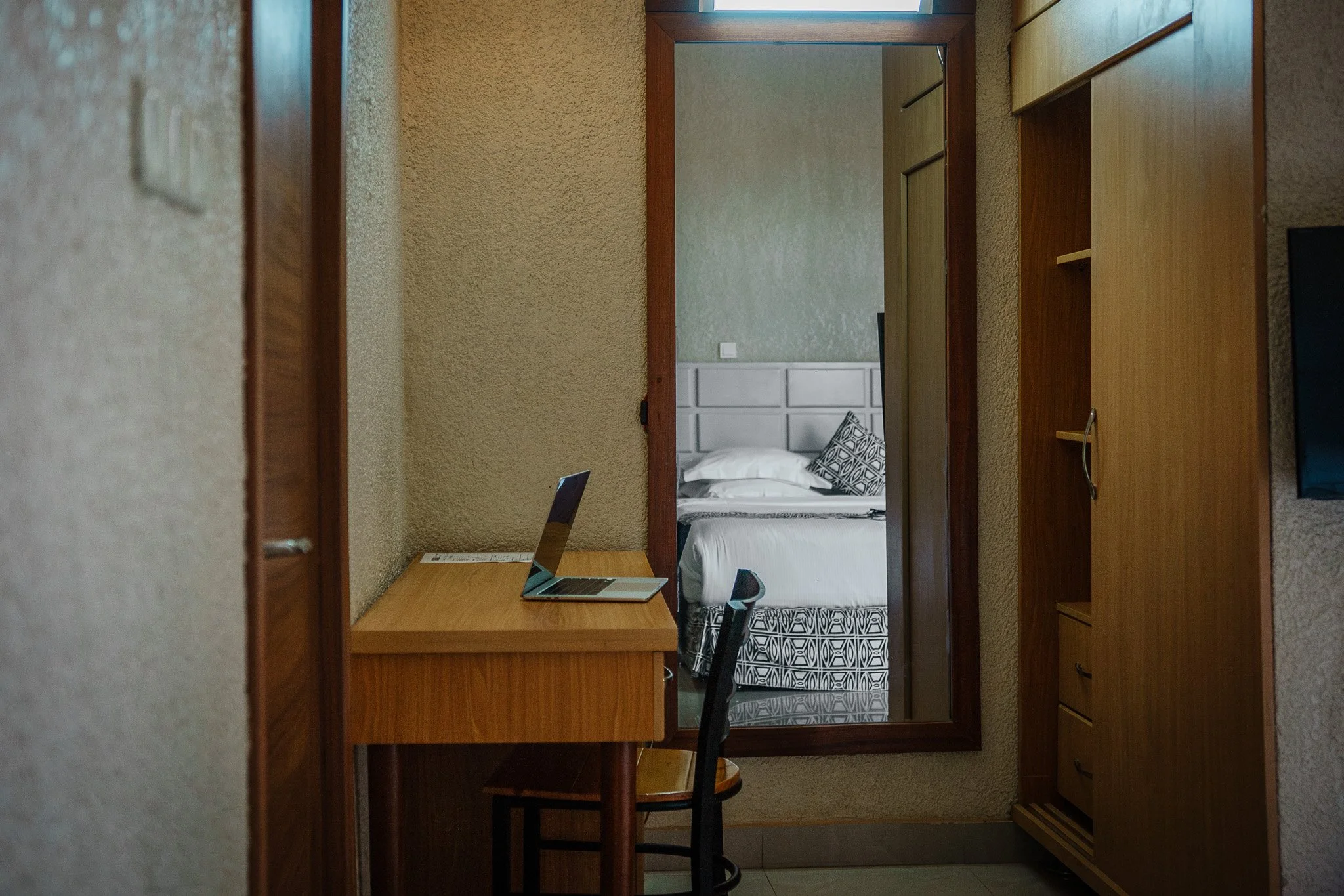 Quiet hotel room at Quality INN Hotel Kigali, Rwanda reflected in a dressing mirror showing bed with white linens and patterned pillow, wooden desk with open laptop, and chair.
