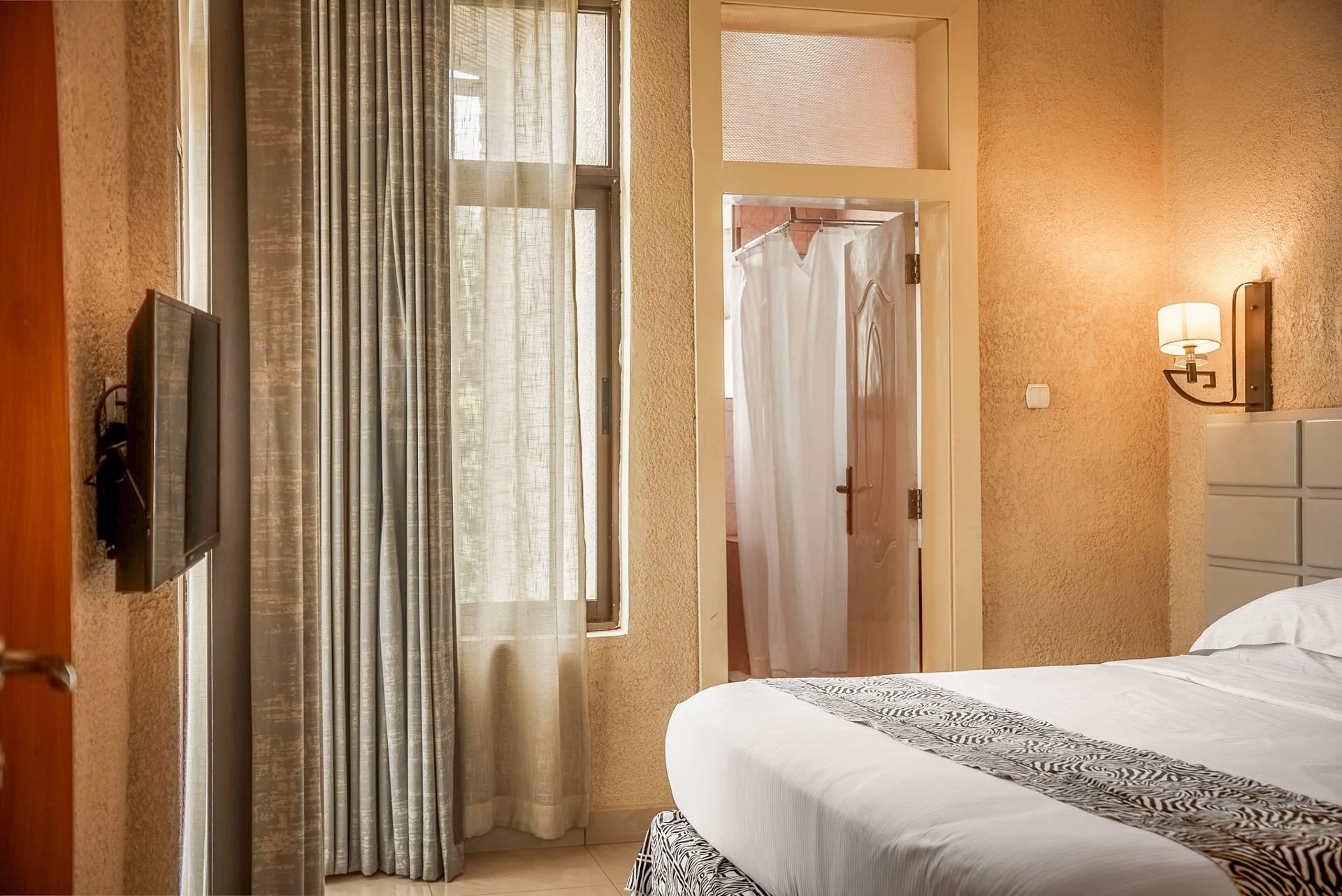 A quiet hotel room at Quality INN Hotel Kigali, Rwanda with bed, wall-mounted TV, window with curtains, and bathroom with shower curtain.