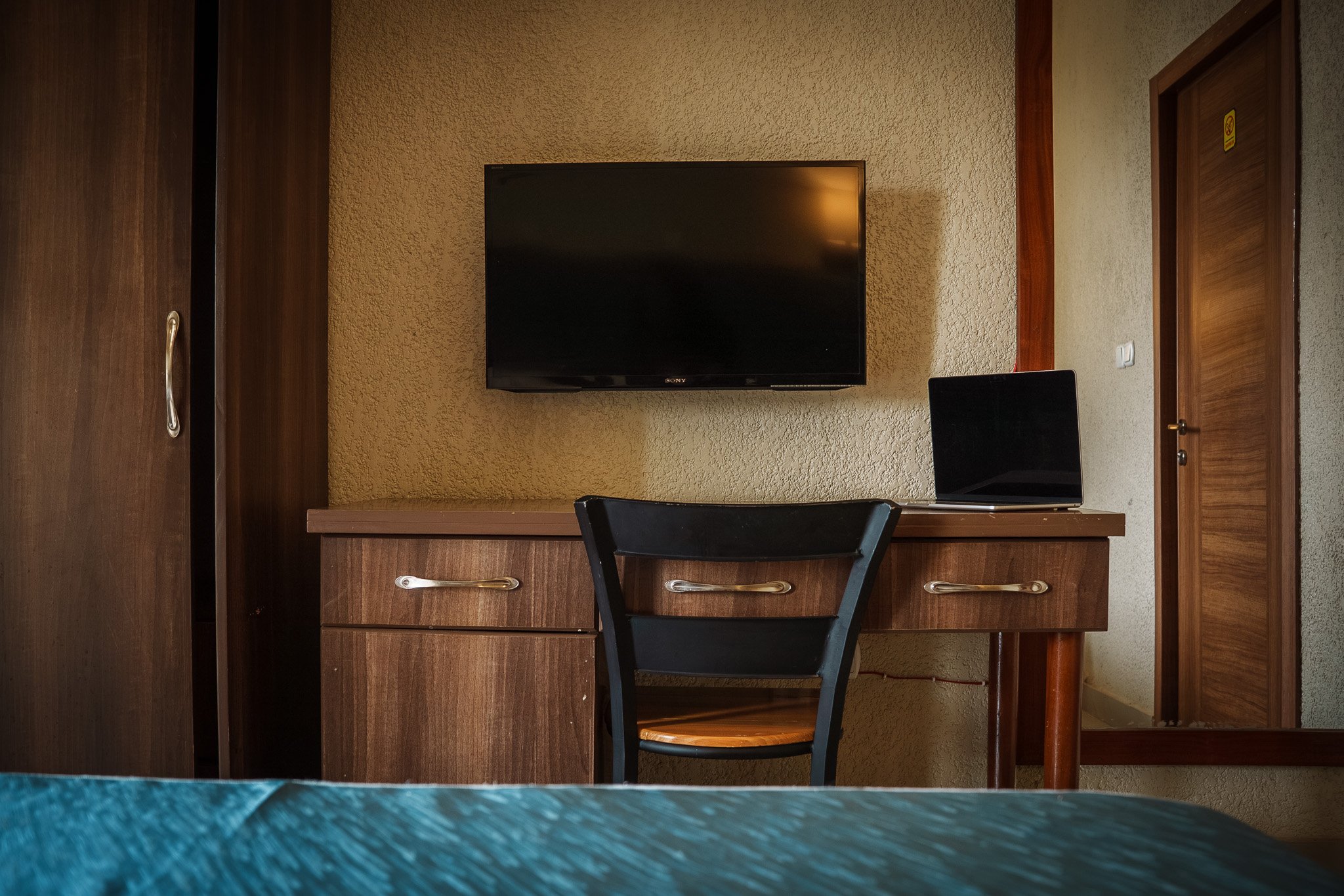 Hotel room at Quality INN Hotel Kigali, Rwanda with wooden desk, black chair, wall-mounted TV, laptop on desk, wardrobe, and mirror reflecting door.
