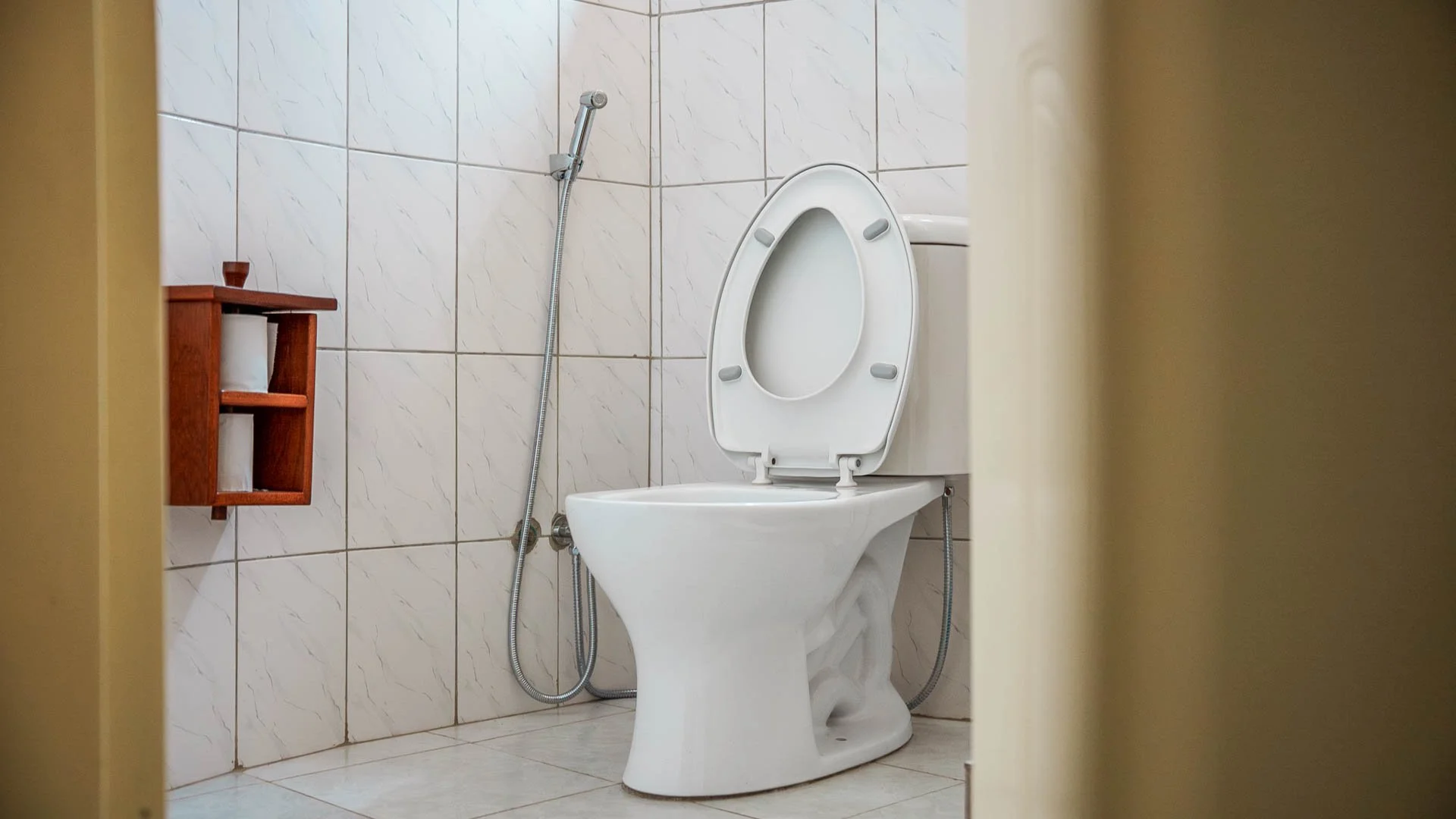 A bathroom with a toilet, a handheld bidet, and a small wooden shelf with toilet paper rolls on it, all against tiled walls at Quality Inn Hotel Kigali, Rwanda.
