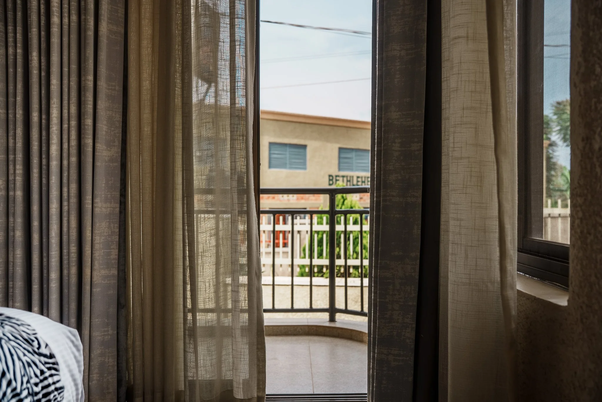View from hotel room at Quality INN Hotel Kigali, Rwanda through partially open curtains to balcony with black railing, overlooking a quiet nearby street.