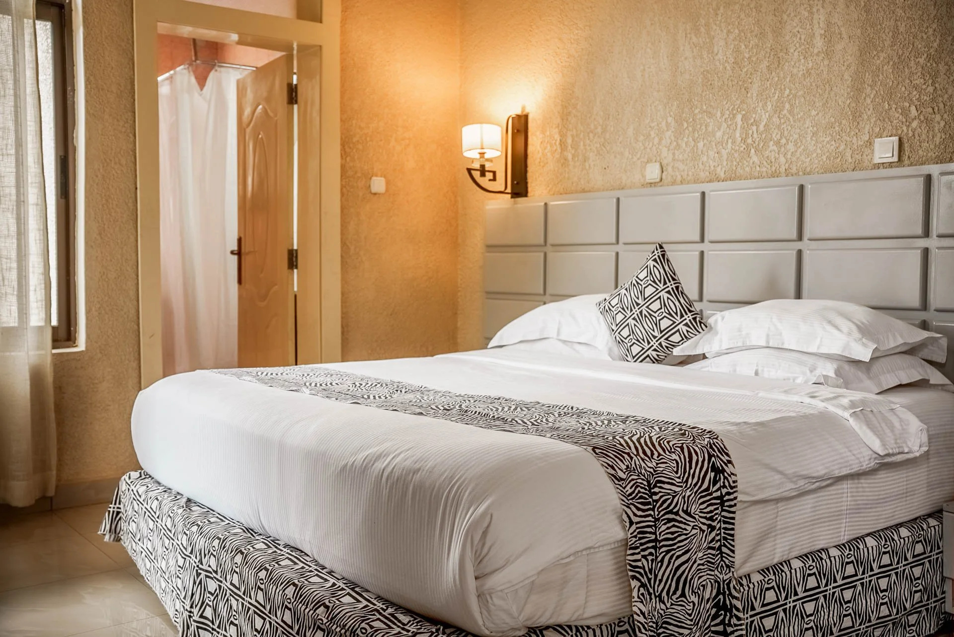 Quiet hotel room at Quality INN Hotel Kigali, Rwanda with large bed, decorative pillows, geometric bed runner, beige walls, wall-mounted lamp, and shower with curtain.