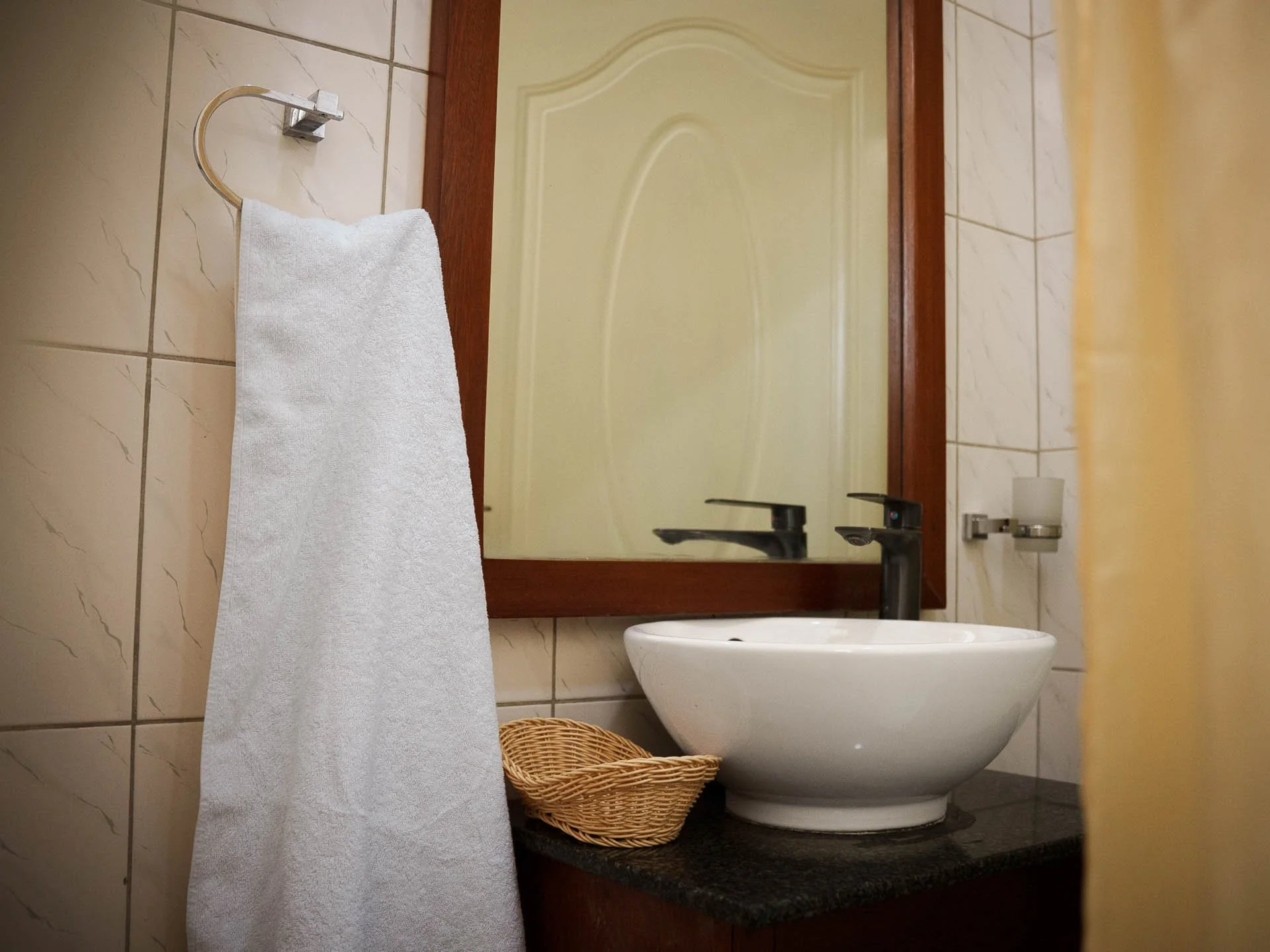 Bathroom countertop with a white circular vessel sink, a black faucet, a small woven basket, a mirror, a tissue holder, a towel hanging on a towel ring, and a yellow shower curtain partially visible on the right at Quality Inn Hotel Kigali, Rwanda.