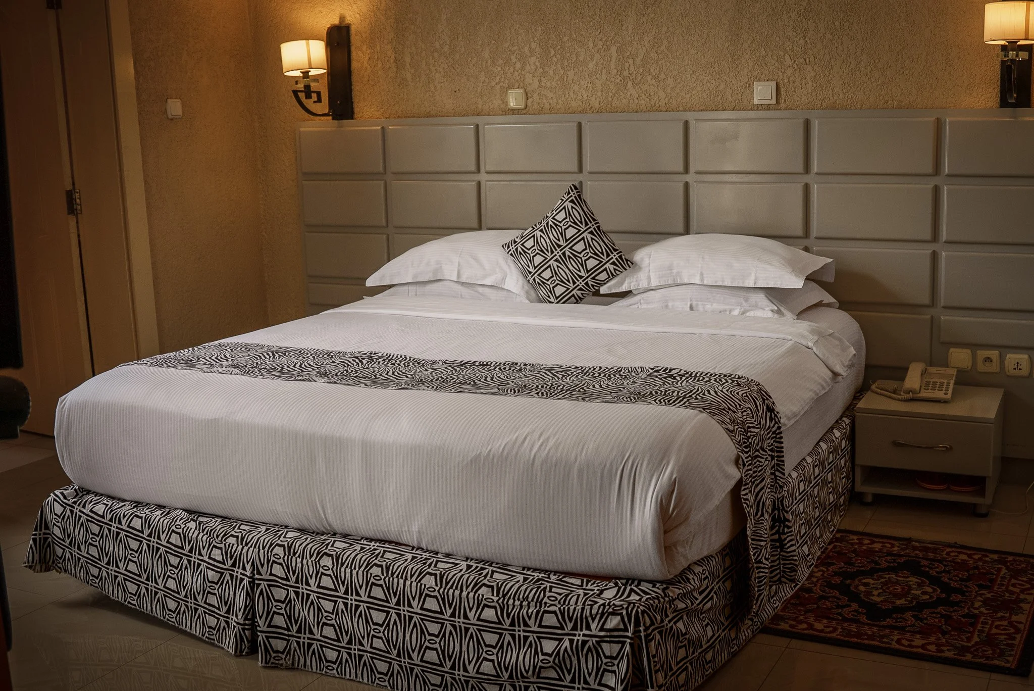 Quiet hotel room at Quality INN Hotel Kigali, Rwanda with bed featuring black and white patterned bedding, wall-mounted lamps, padded headboard, bedside table with telephone, and small floor rug.