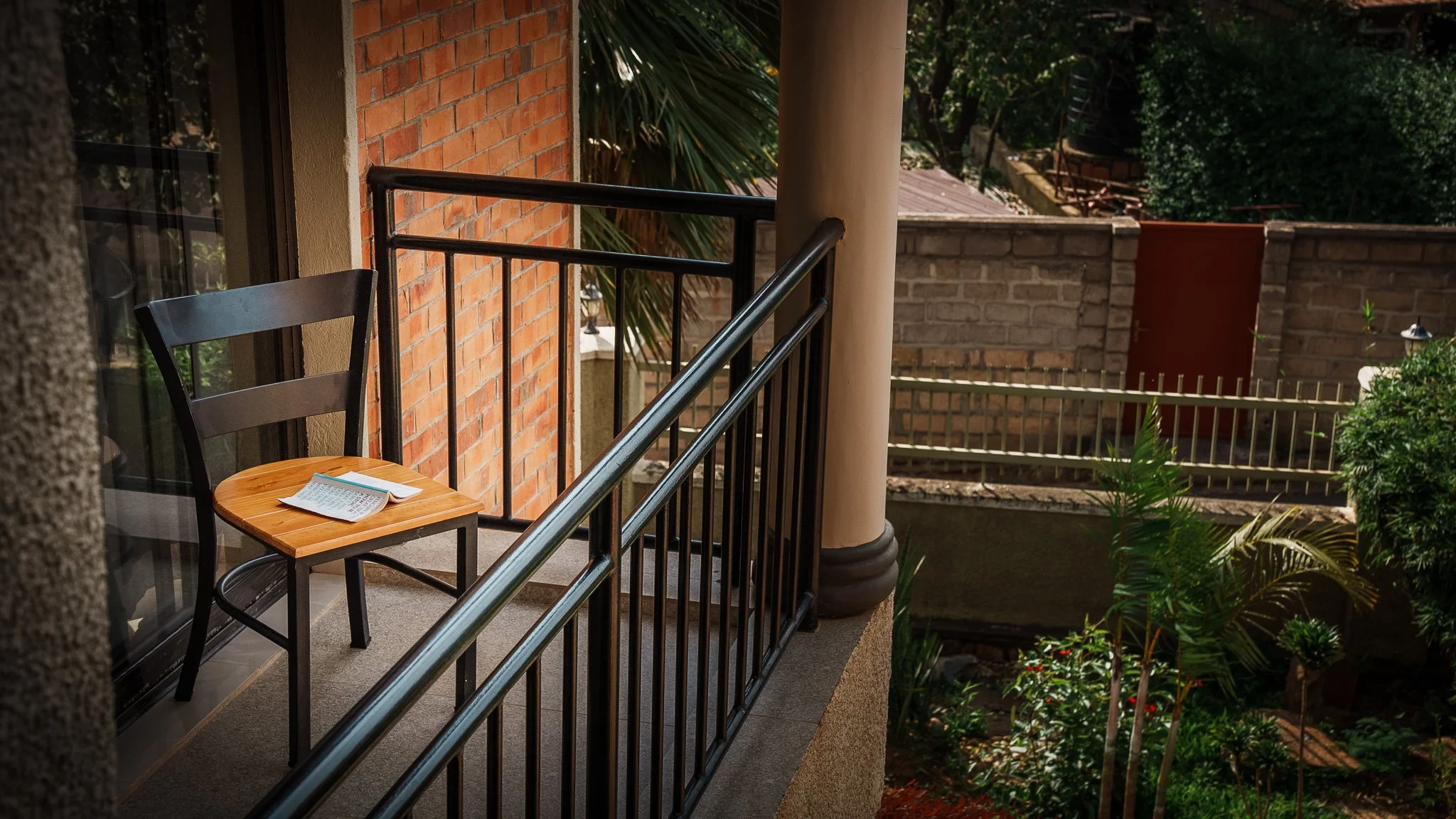 Balcony at Quality INN Hotel Kigali, Rwanda with wooden chair and book, overlooking greenery and the nearby neighborhood.