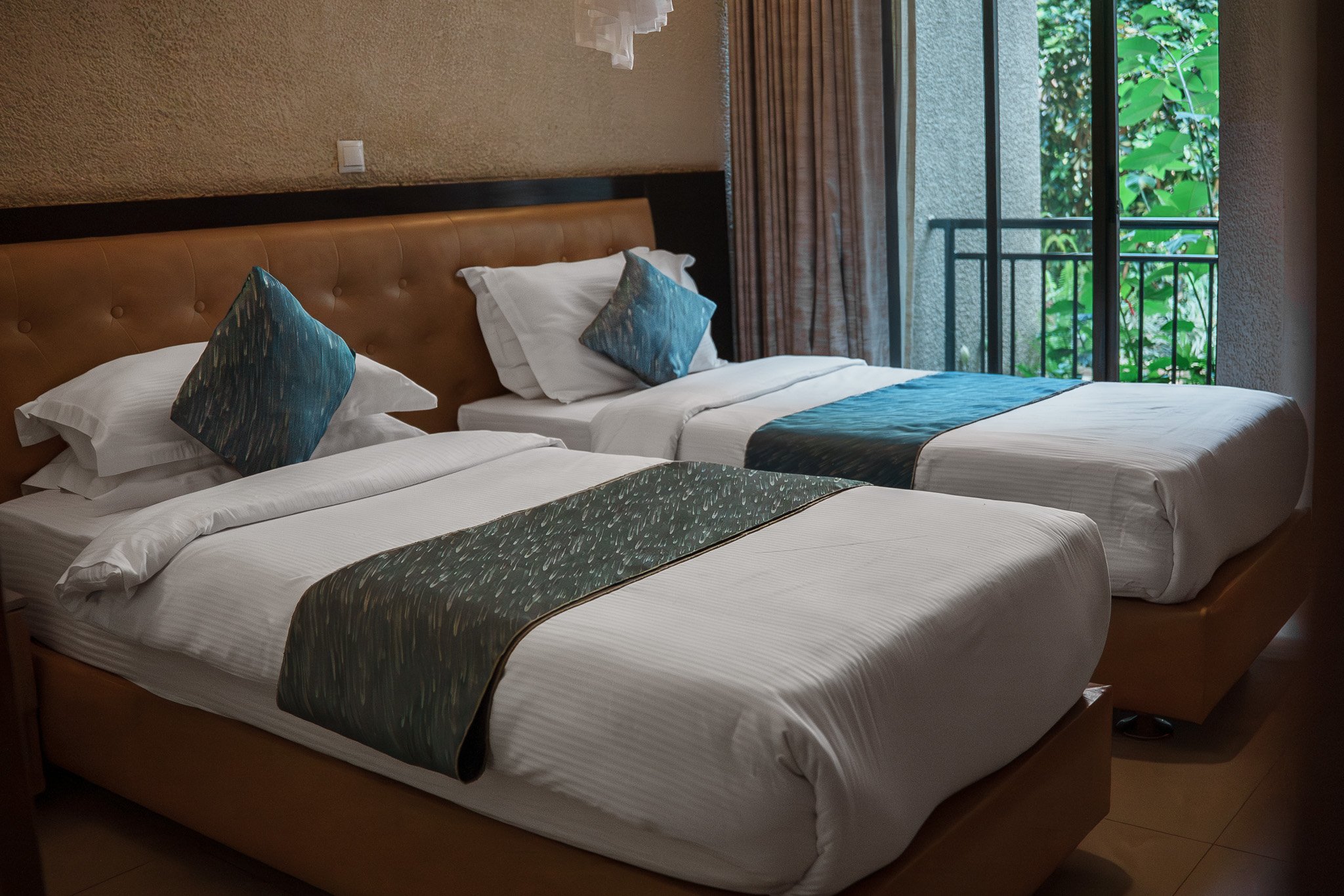 Twin bedroom at Quality INN Hotel Kigali, Rwanda with white linens, blue and green bed runners, and balcony overlooking greenery.