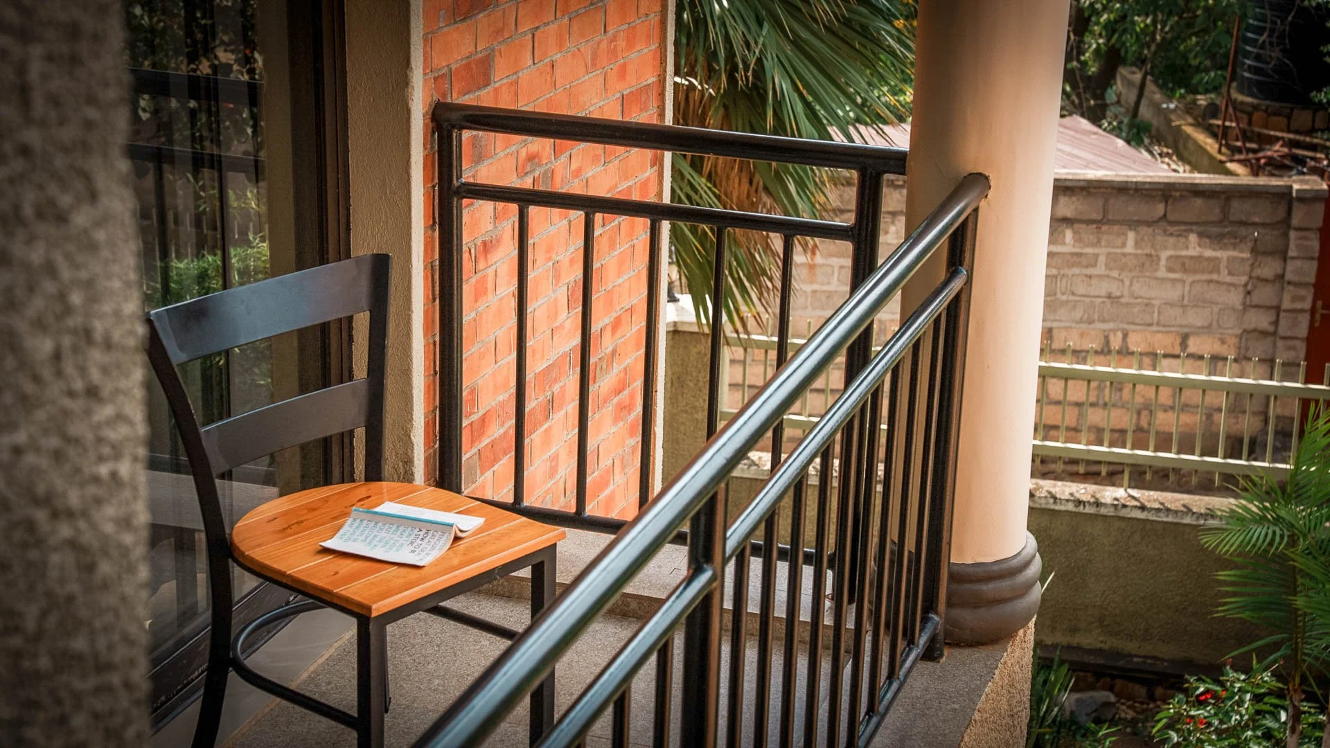 Balcony at Quality INN Hotel Kigali, Rwanda with wooden chair and book, overlooking greenery and the nearby neighborhood.