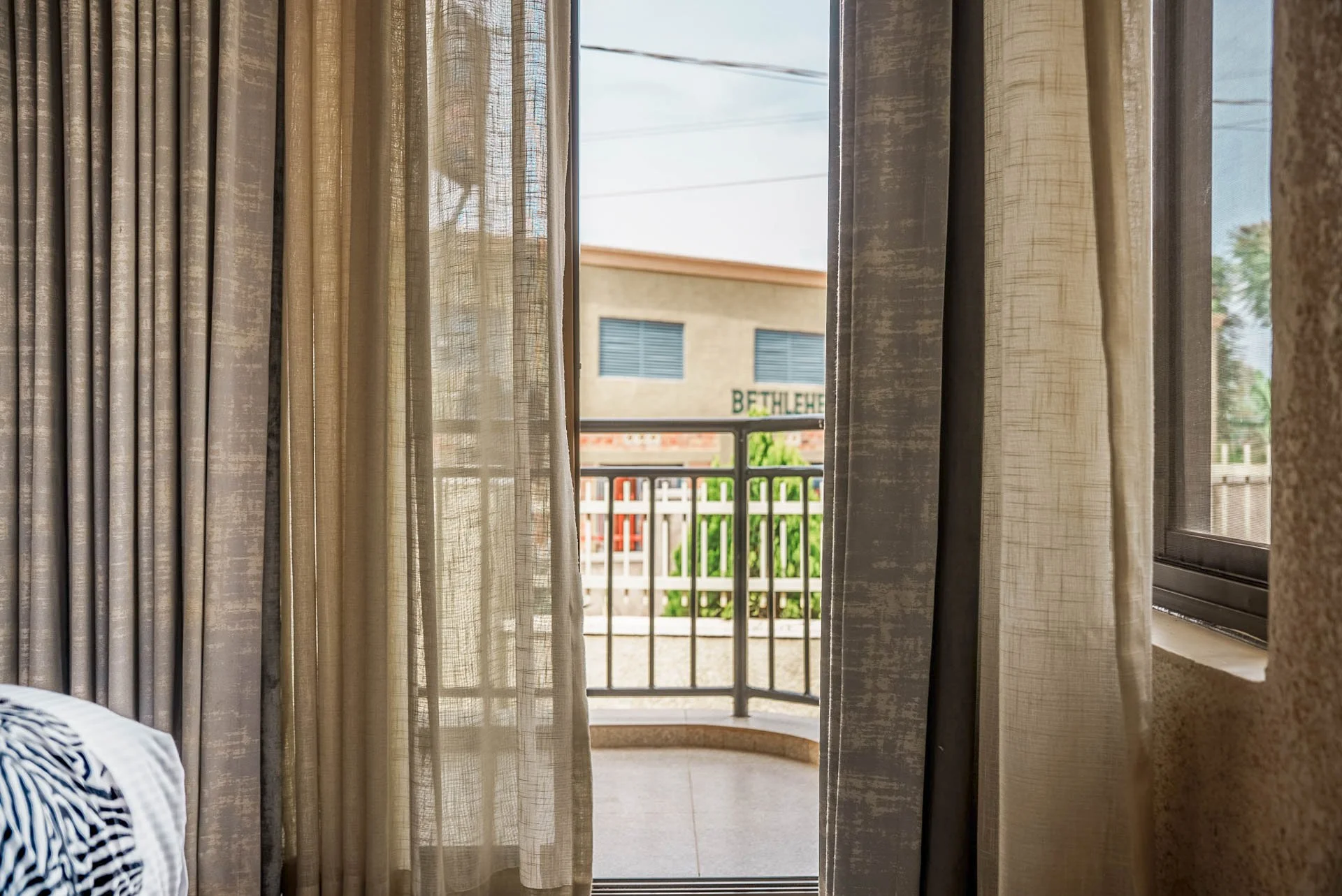 View from hotel room at Quality INN Hotel Kigali, Rwanda through partially open curtains to balcony with black railing, overlooking a quiet nearby street.