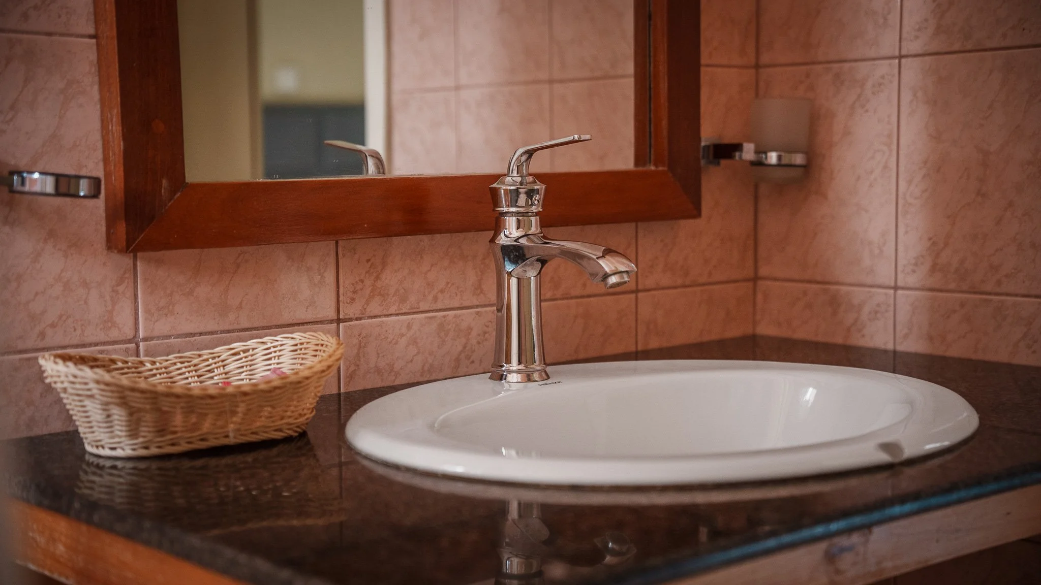 Bathroom sink with a chrome faucet, a mirror above, a small wicker basket to the left, pink tile walls, and a dark countertop at Quality Inn Hotel Kigali, Rwanda