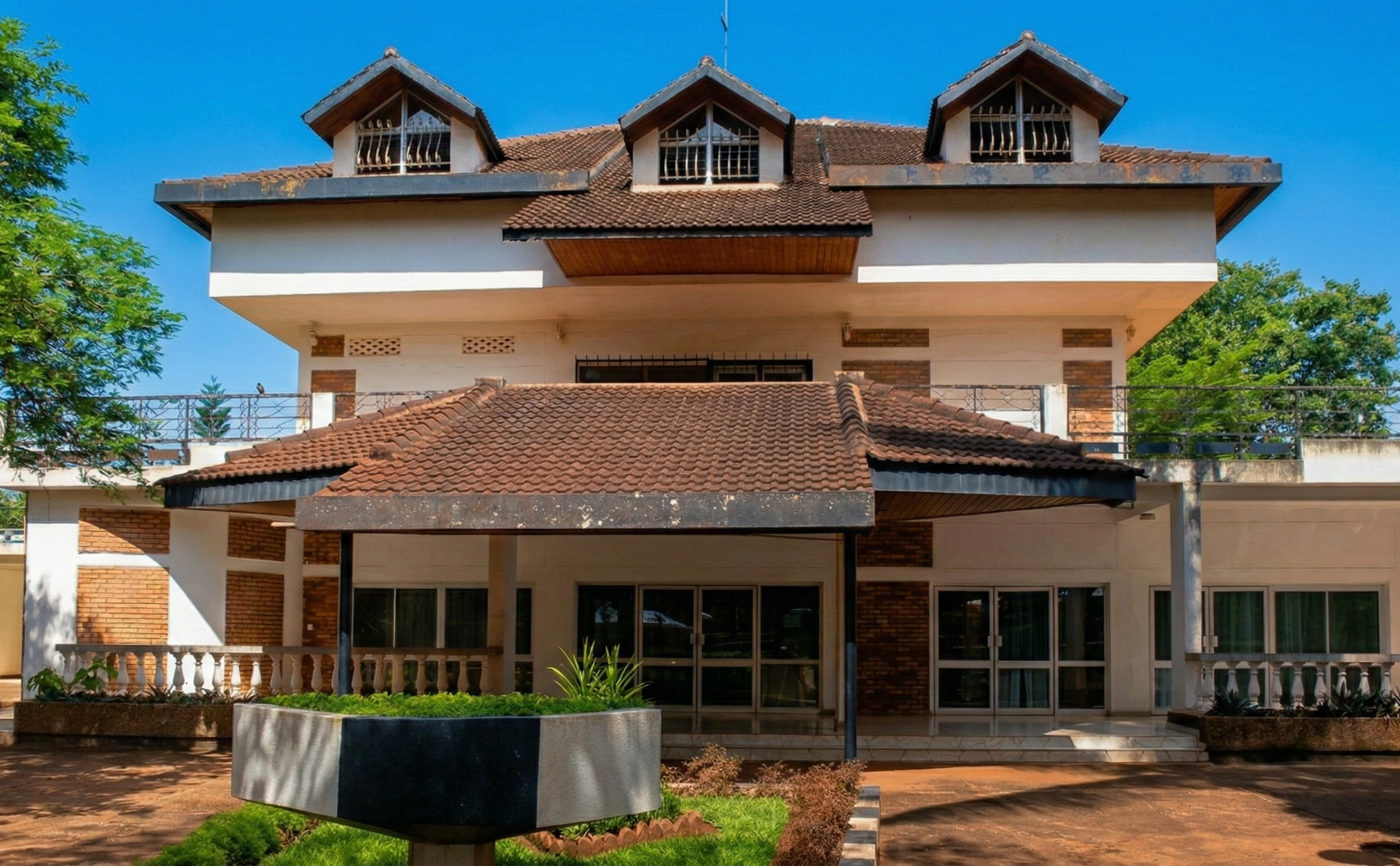 Rwanda Art Museum, two stories with white walls, brick accents, and a brown tiled roof. Three dormer windows on top, a dark-tiled canopy over the entrance, and a paved courtyard with a stone planter and white balustrades.