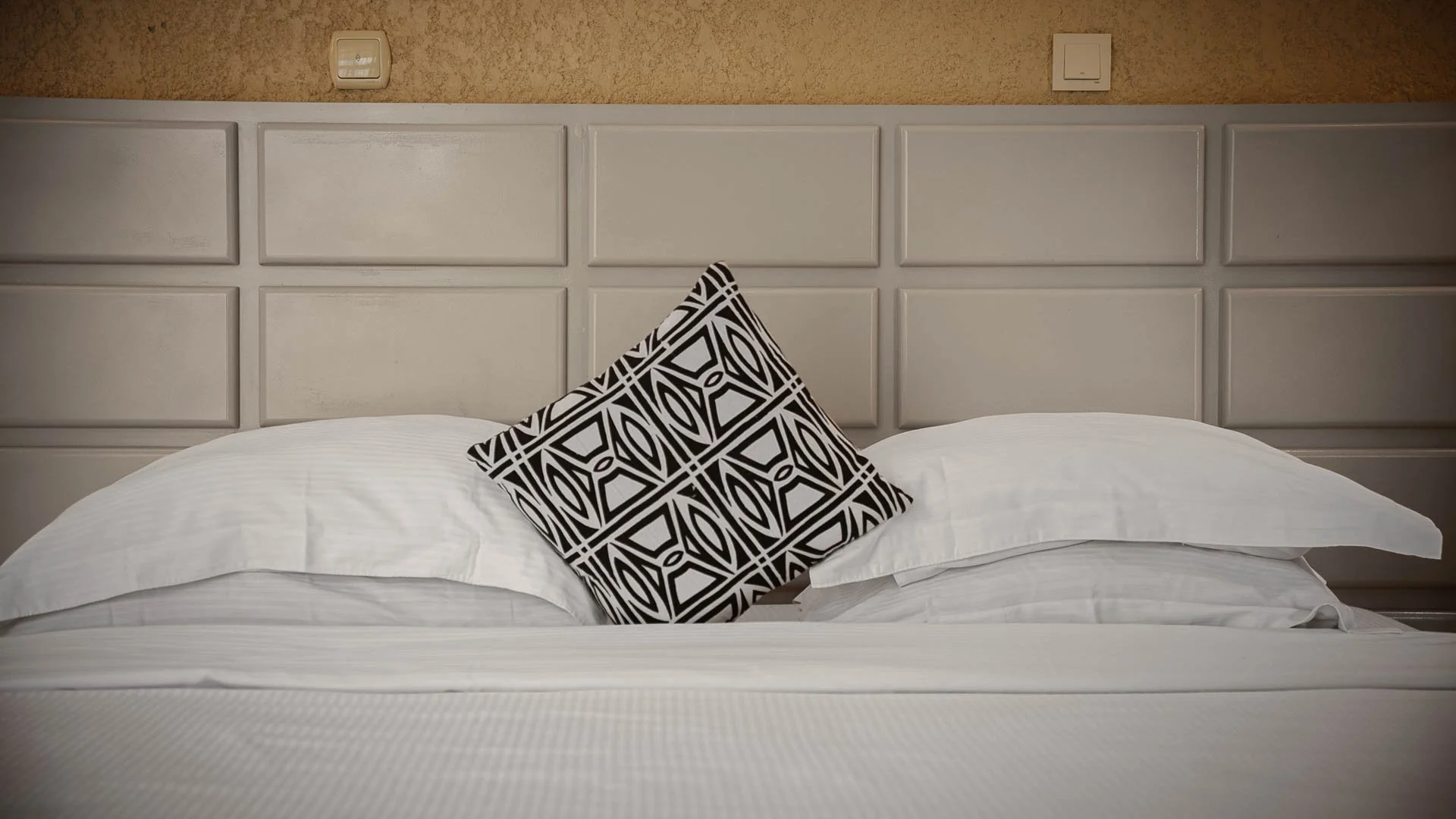 A neatly made bed with white bed linens and a decorative black-and-white patterned pillow. The bed has a beige, paneled headboard and appears to be in a hotel room.