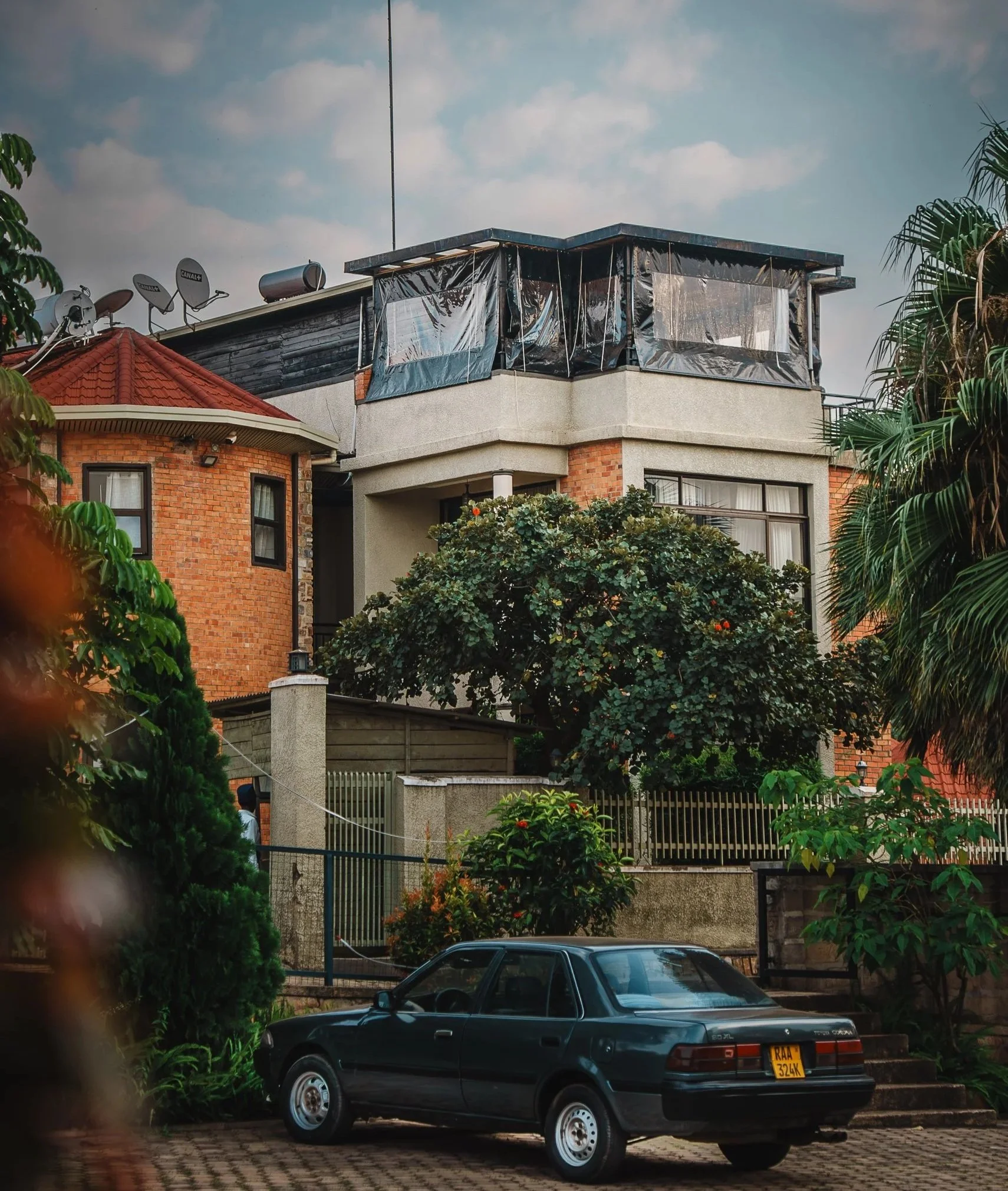Two-story property at Quality INN Hotel Kigali, Rwanda with brick and concrete exterior, tree in front, solar panels and satellite dishes on the roof, dark green car parked in the driveway, and rooftop lounge on the right side of the building.