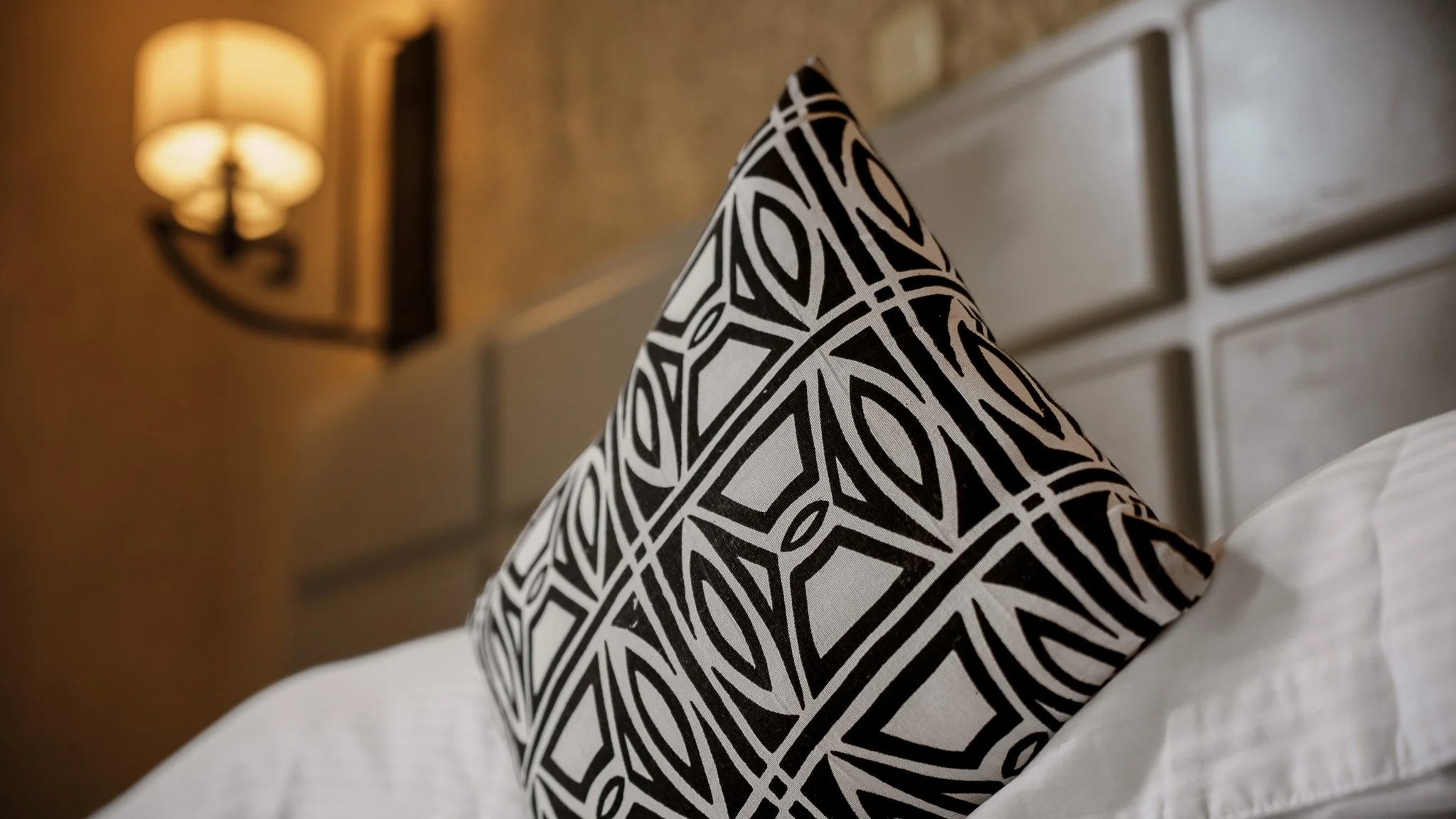 Decorative black and white patterned pillow on a bed with white sheets at Quality INN Hotel Kigali, Rwanda, with wall-mounted lamp in the background.