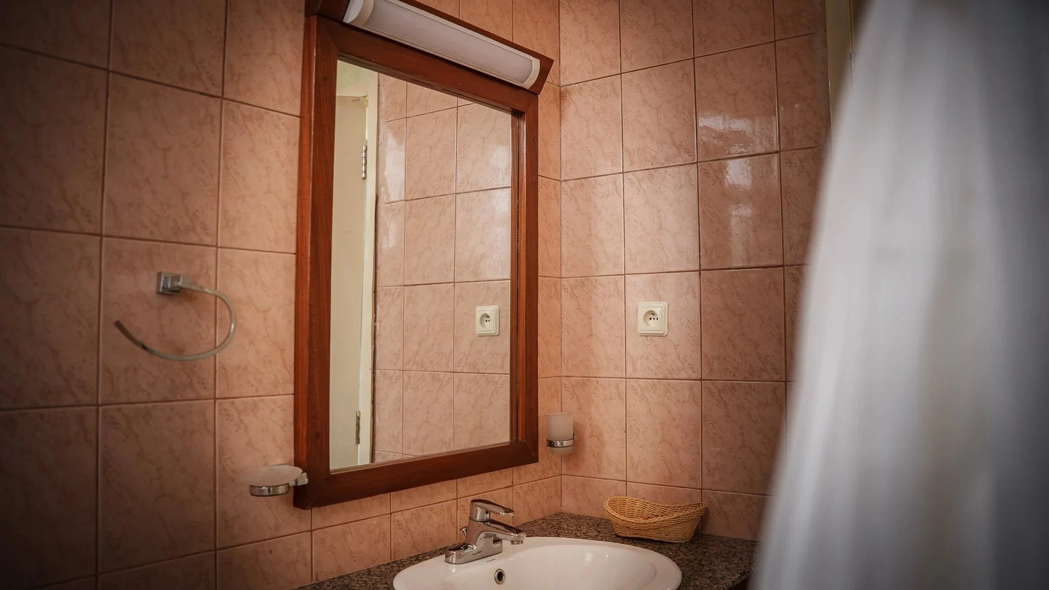 Bathroom mirror with wooden frame, pink tiled wall, white sink with granite countertop, wicker basket, wall-mounted soap dispenser, electrical outlets, and a partial view of a shower curtain at Quality Inn Hotel Kigali, Rwanda.