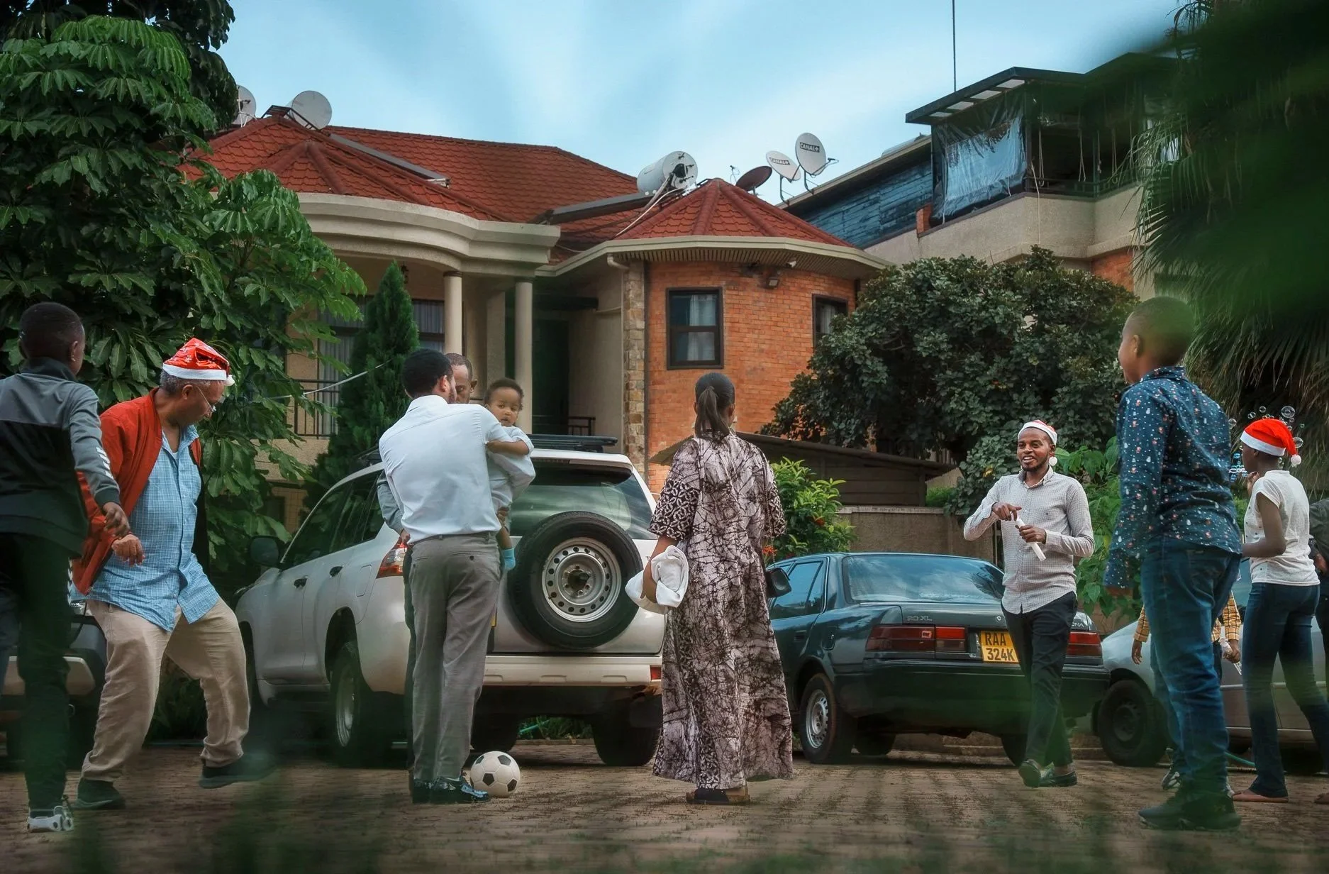 Group of people celebrating outdoors in front of  Quality INN Hotel Kigali, Rwanda with some wearing Santa hats, a man dressed as Santa, and children playing with a soccer ball.