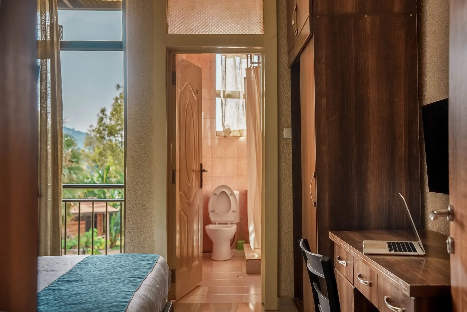 Hotel room at Quality INN Hotel Kigali, Rwanda with bed, open balcony door, wall-mounted TV, work desk, wardrobe, and bathroom with toilet and window.