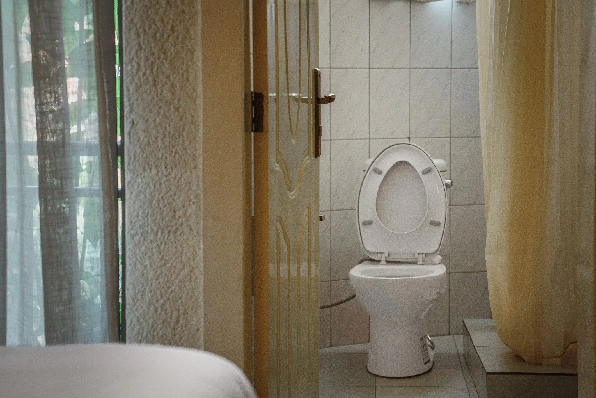 View of a bathroom with a white toilet, a partially open door, a yellow shower curtain, and a window with curtains at Quality Inn Hotel Kigali, Rwanda.