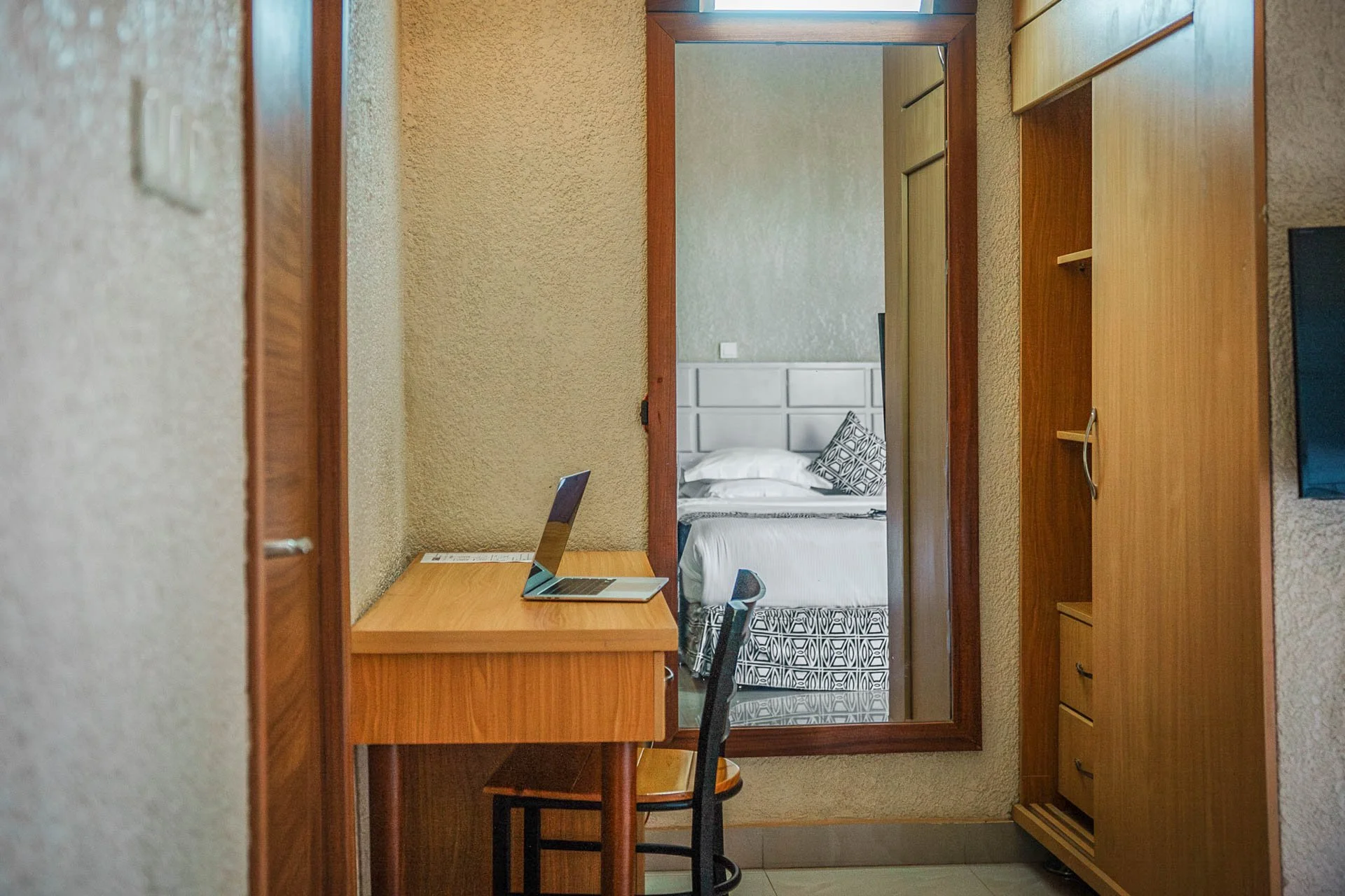 Quiet hotel room at Quality INN Hotel Kigali, Rwanda reflected in a dressing mirror showing bed with white linens and patterned pillow, wooden desk with open laptop, and chair.