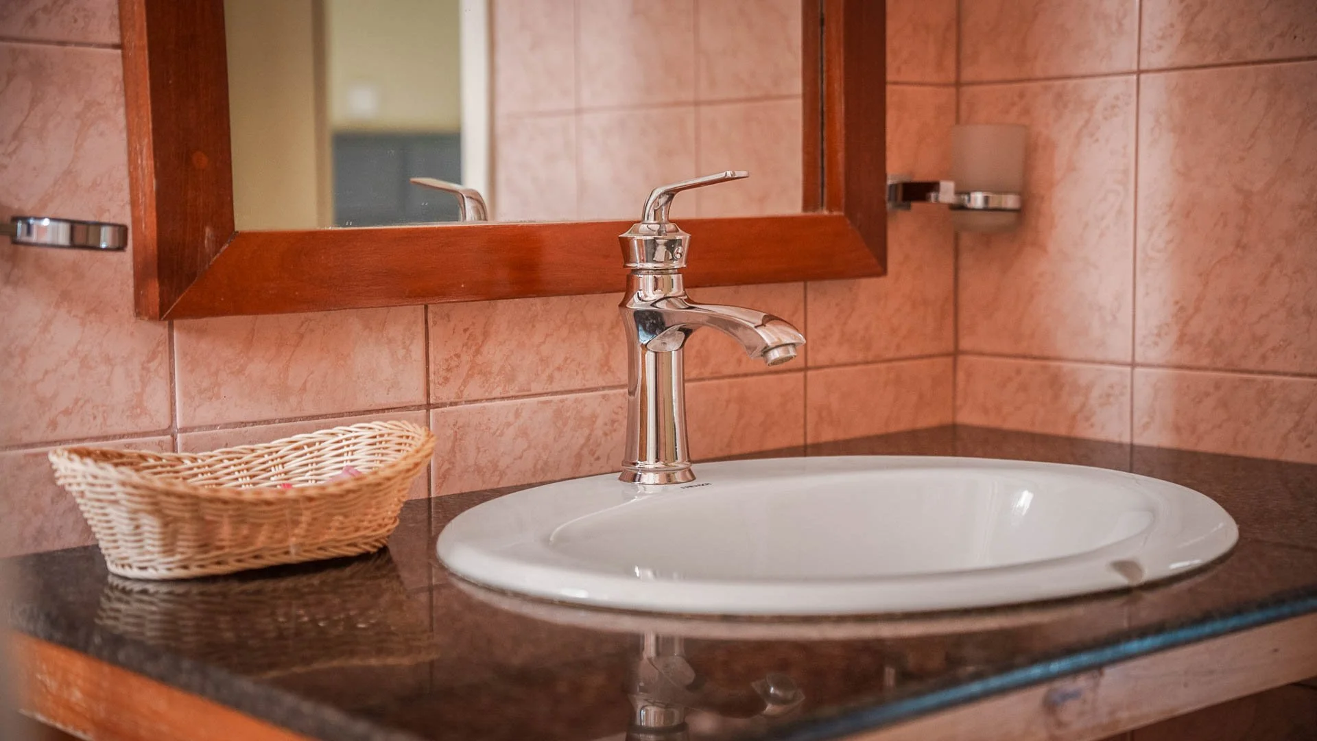 Bathroom sink with a chrome faucet, a mirror above, a small wicker basket to the left, pink tile walls, and a dark countertop at Quality Inn Hotel Kigali, Rwanda