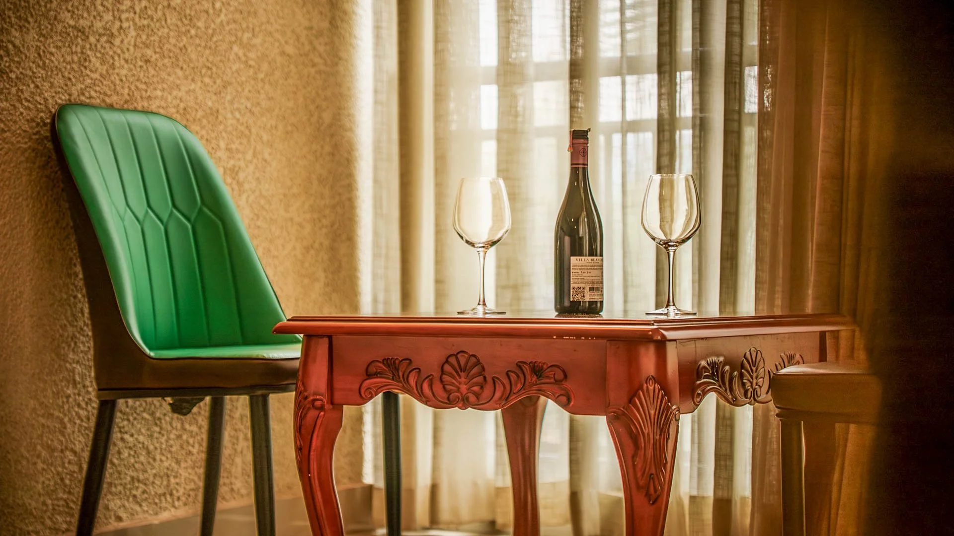 Wine bottle and glasses on a carved wooden table next to a large window in the King Studio at Quality Inn Hotel Kigali, Rwanda—a welcoming spot for couples and friends to relax.