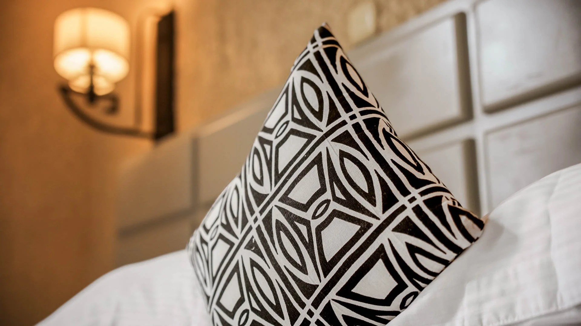 Decorative black and white patterned pillow on a bed with white sheets at Quality INN Hotel Kigali, Rwanda, with wall-mounted lamp in the background.