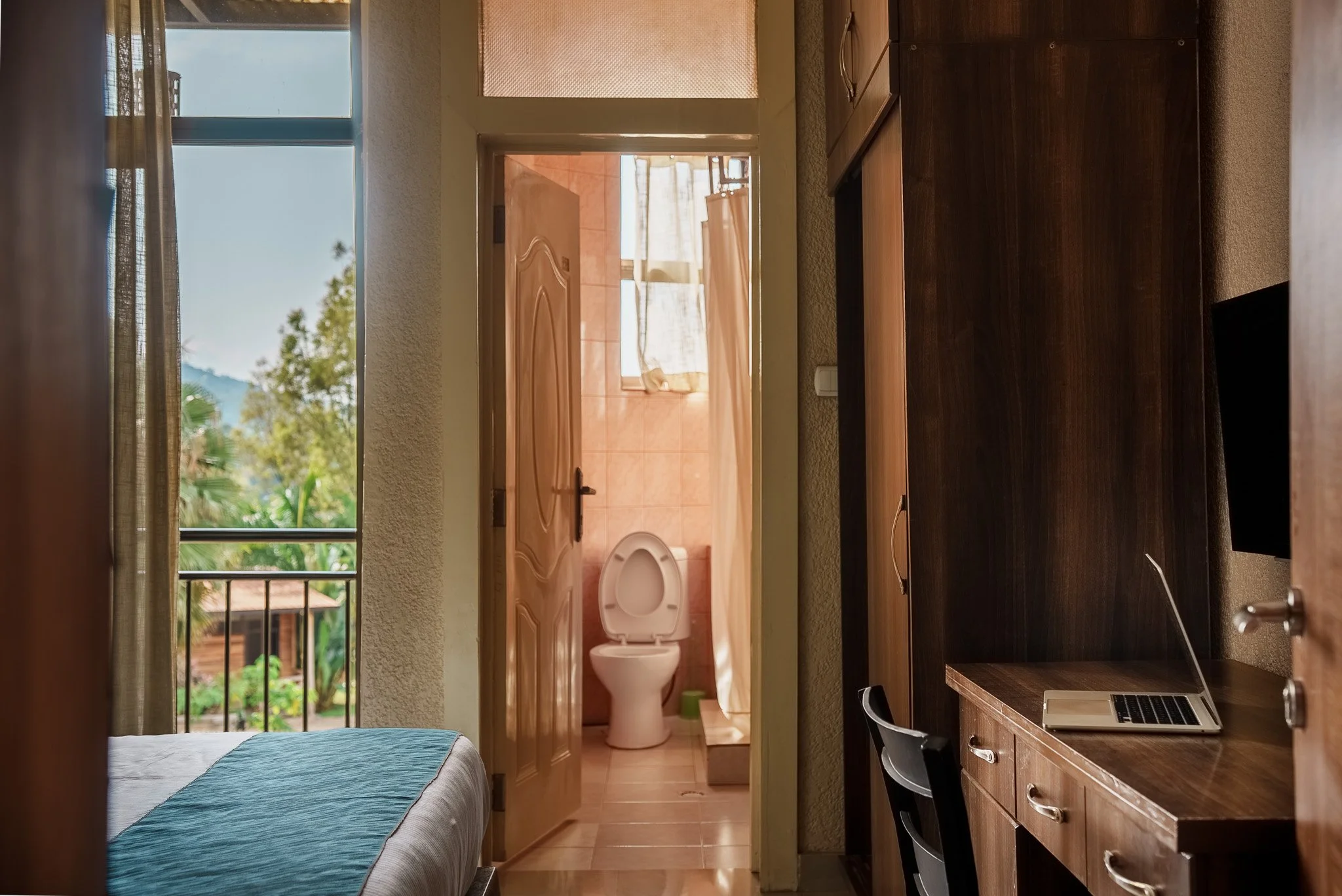 Hotel room at Quality INN Hotel Kigali, Rwanda with bed, open balcony door, wall-mounted TV, work desk, wardrobe, and bathroom with toilet and window.