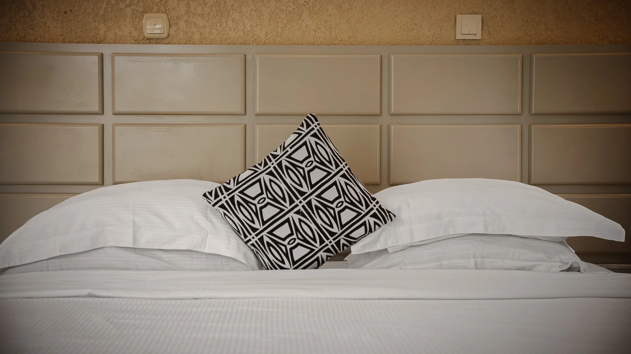 A neatly made bed with white bed linens and a decorative black-and-white patterned pillow. The bed has a beige, paneled headboard and appears to be in a hotel room.