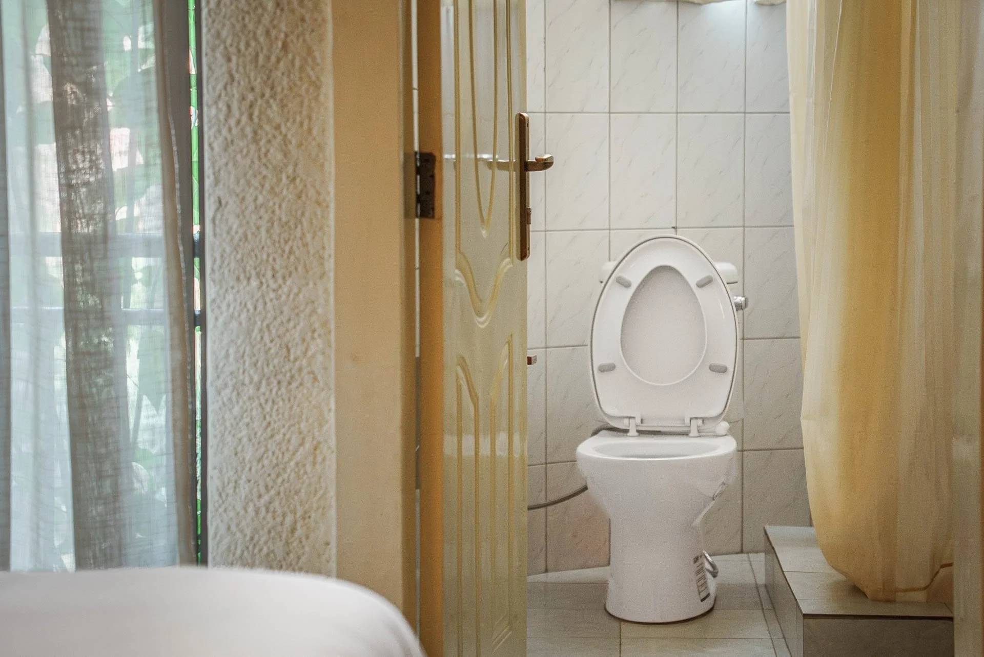 View of a bathroom with a white toilet, a partially open door, a yellow shower curtain, and a window with curtains at Quality Inn Hotel Kigali, Rwanda.