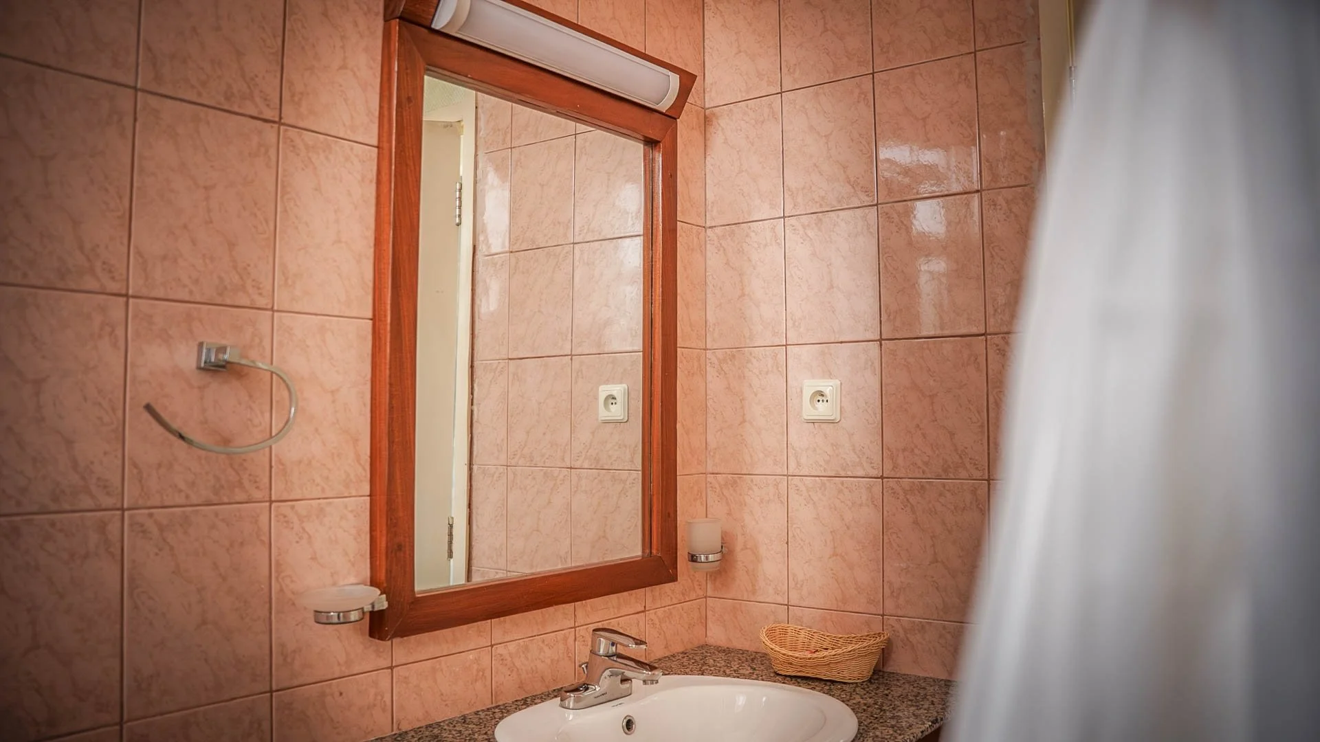 Bathroom mirror with wooden frame, pink tiled wall, white sink with granite countertop, wicker basket, wall-mounted soap dispenser, electrical outlets, and a partial view of a shower curtain at Quality Inn Hotel Kigali, Rwanda.