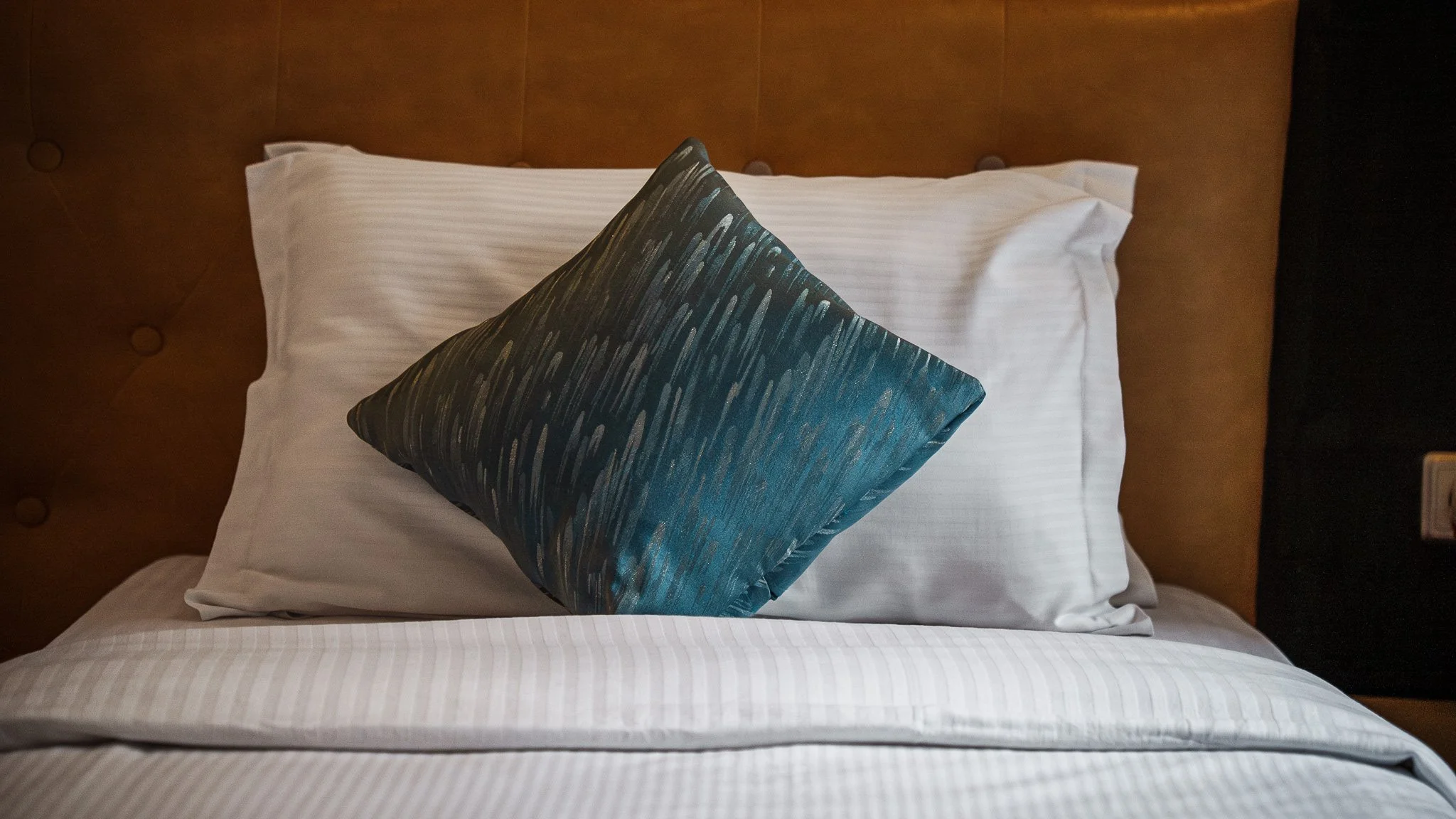Neatly made bed inside Deluxe Twin Room at Quality INN Hotel Kigali, Rwanda with white pillow and decorative dark blue pillow against brown headboard.