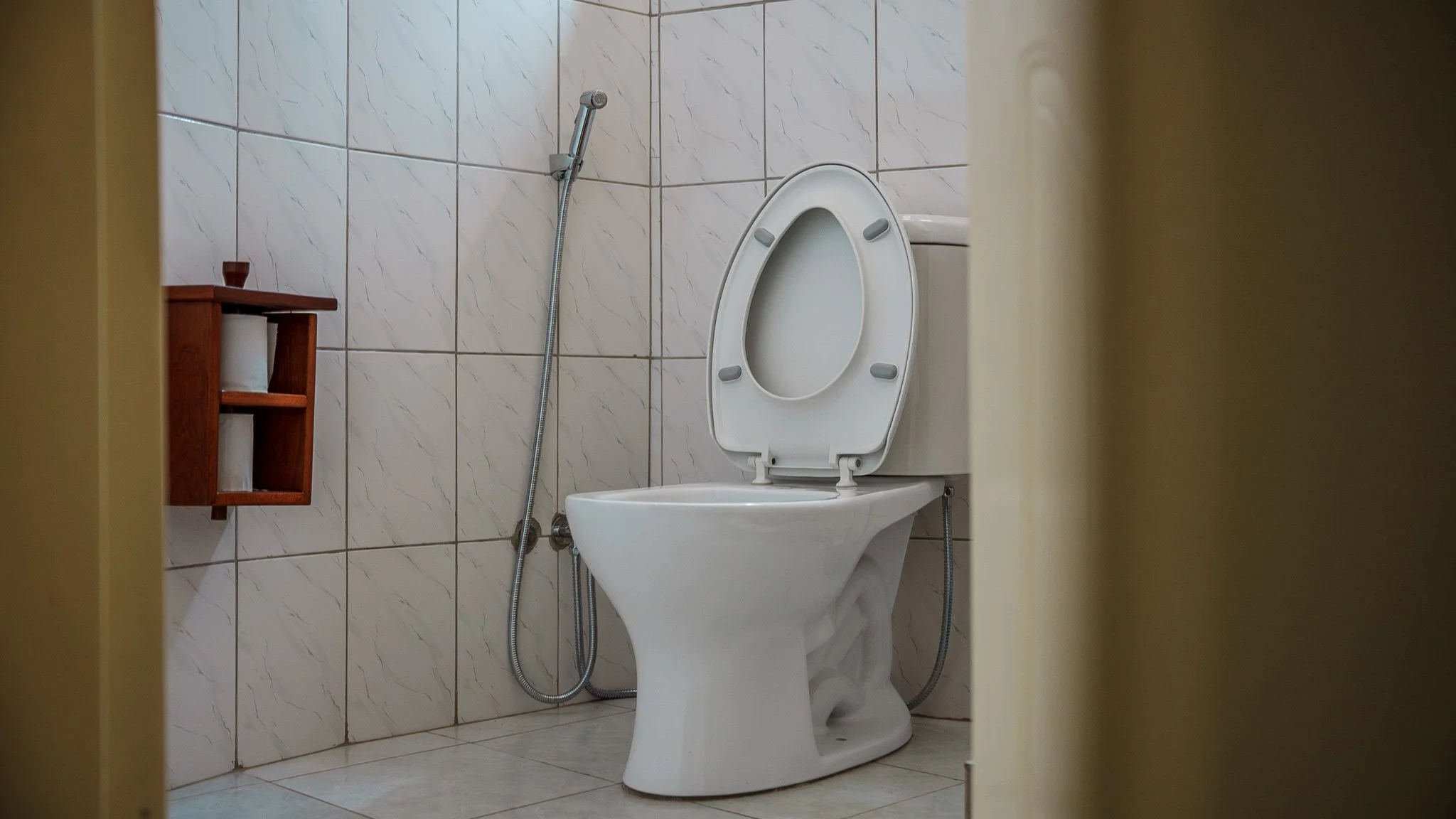 A bathroom with a toilet, a handheld bidet, and a small wooden shelf with toilet paper rolls on it, all against tiled walls at Quality Inn Hotel Kigali, Rwanda.