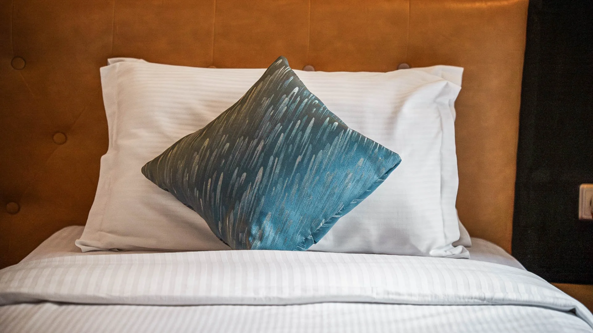 Neatly made bed inside Deluxe Twin Room at Quality INN Hotel Kigali, Rwanda with white pillow and decorative dark blue pillow against brown headboard.