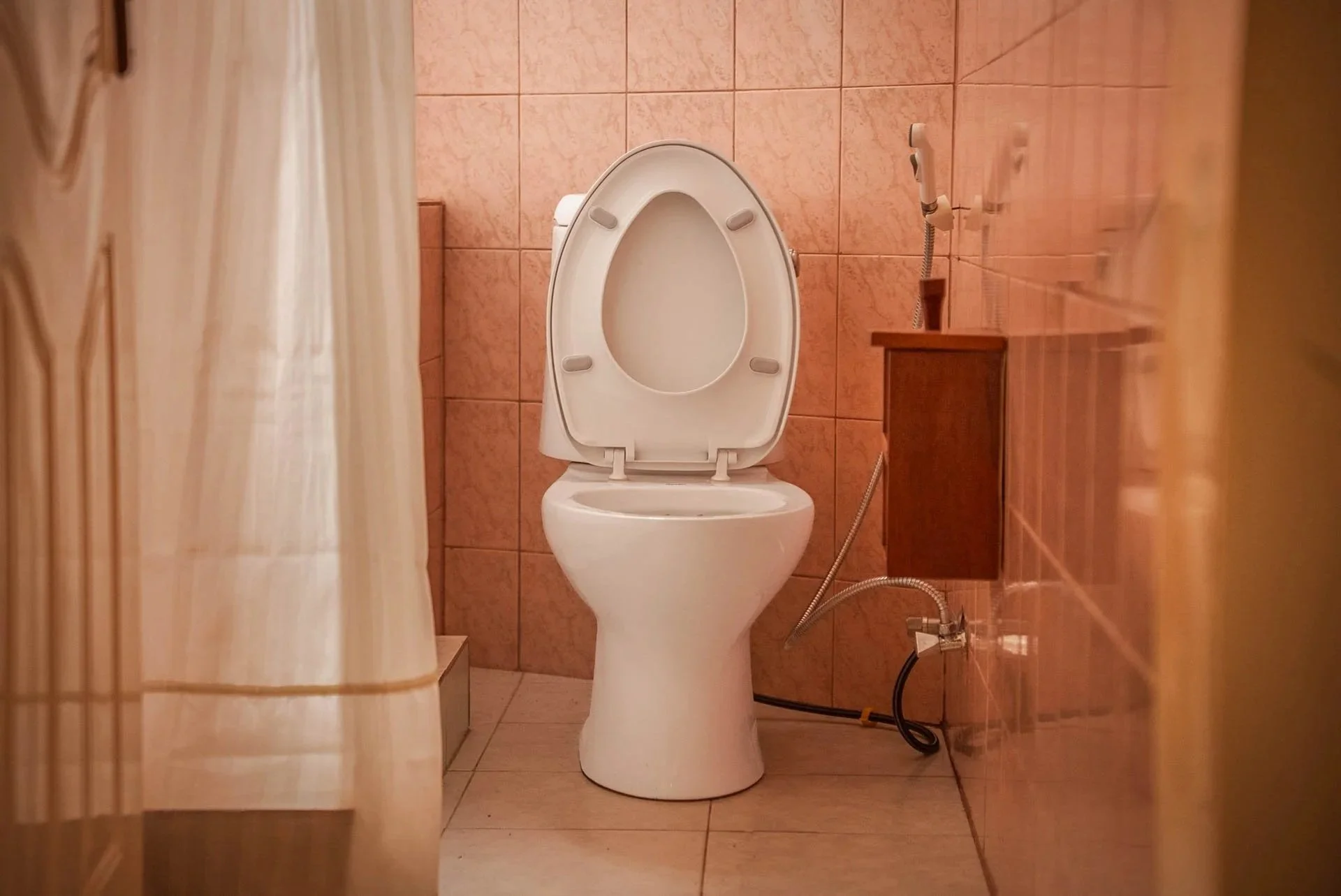 A white toilet with the lid up in a small, tiled bathroom, with a beige curtain partially visible on the left and a wooden shelf or cabinet on the right at Quality Inn Hotel Kigali, Rwanda.