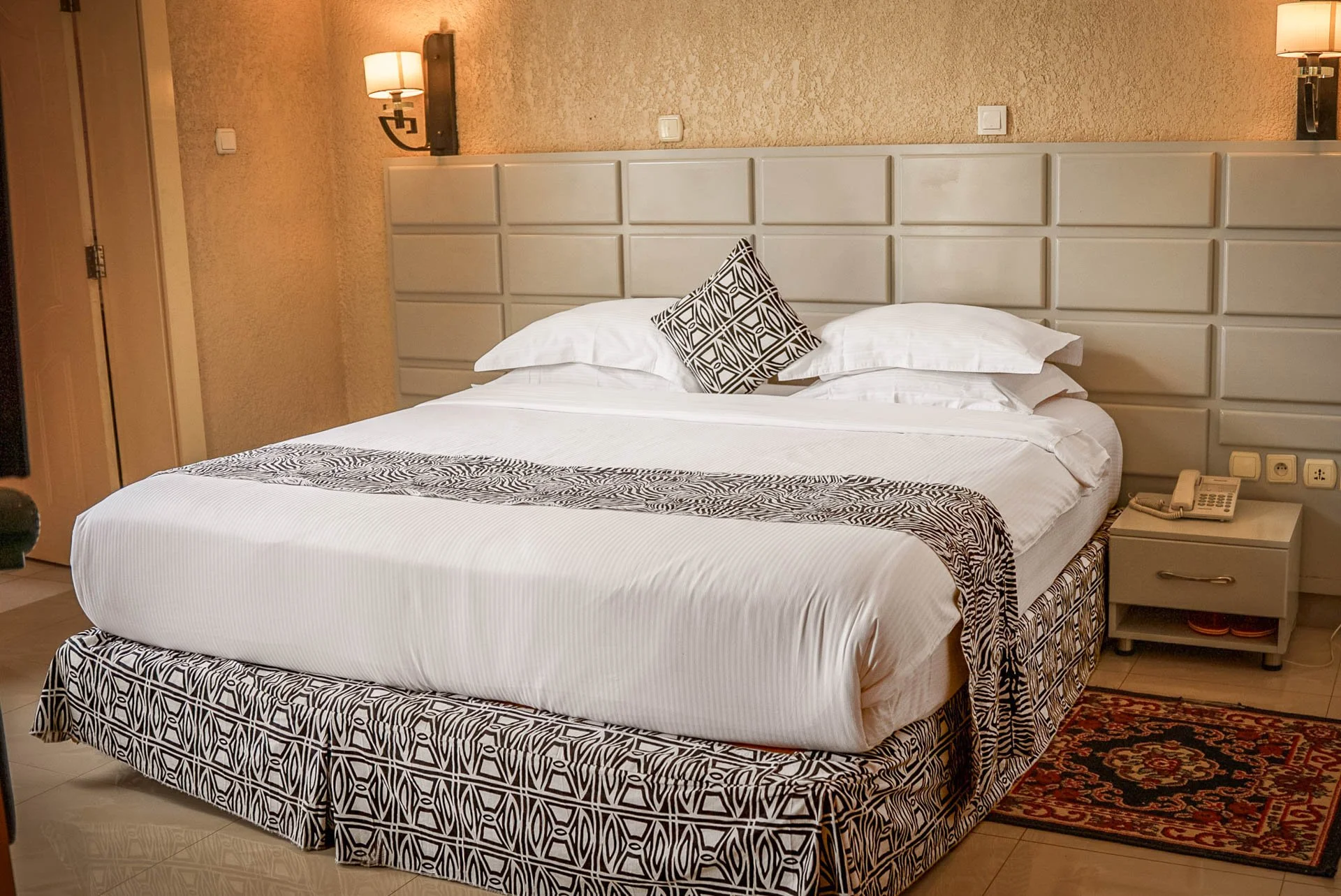 Quiet hotel room at Quality INN Hotel Kigali, Rwanda with bed featuring black and white patterned bedding, wall-mounted lamps, padded headboard, bedside table with telephone, and small floor rug.