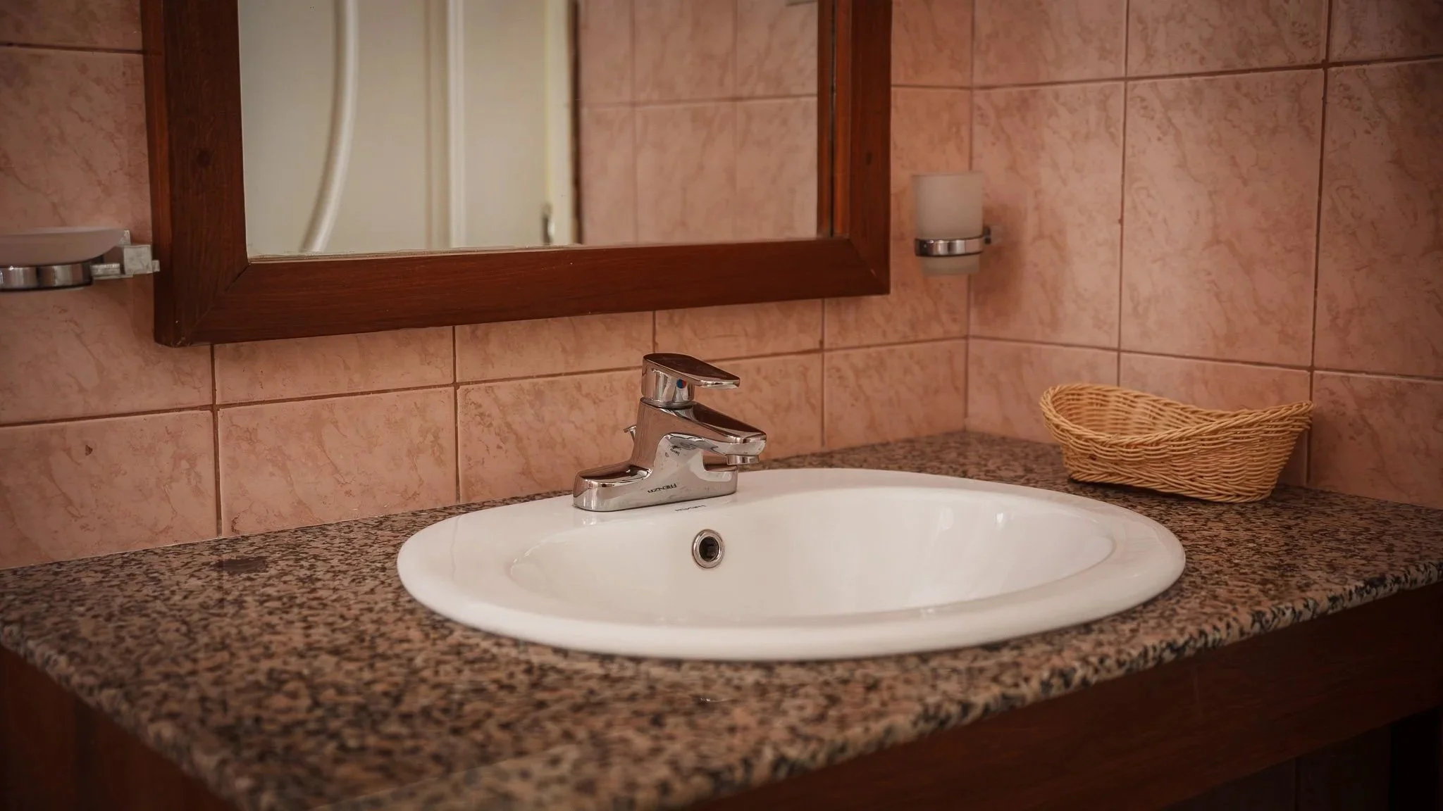 A bathroom sink with a chrome faucet, a granite countertop, a wicker basket, pink tiled wall, a framed mirror, and a wall-mounted light fixture at Quality Inn Hotel Kigali, Rwanda
