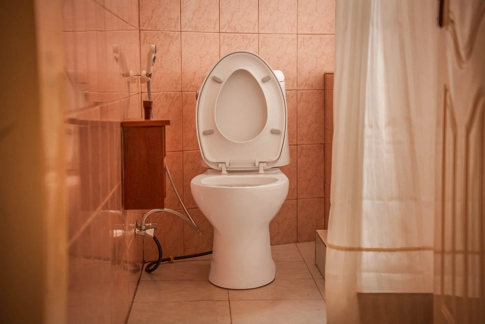 A white toilet with the lid open in a restroom with pinkish-brown tiled walls and floor, partially obscured by a beige shower curtain on the right side at Quality Inn Hotel Kigali, Rwanda