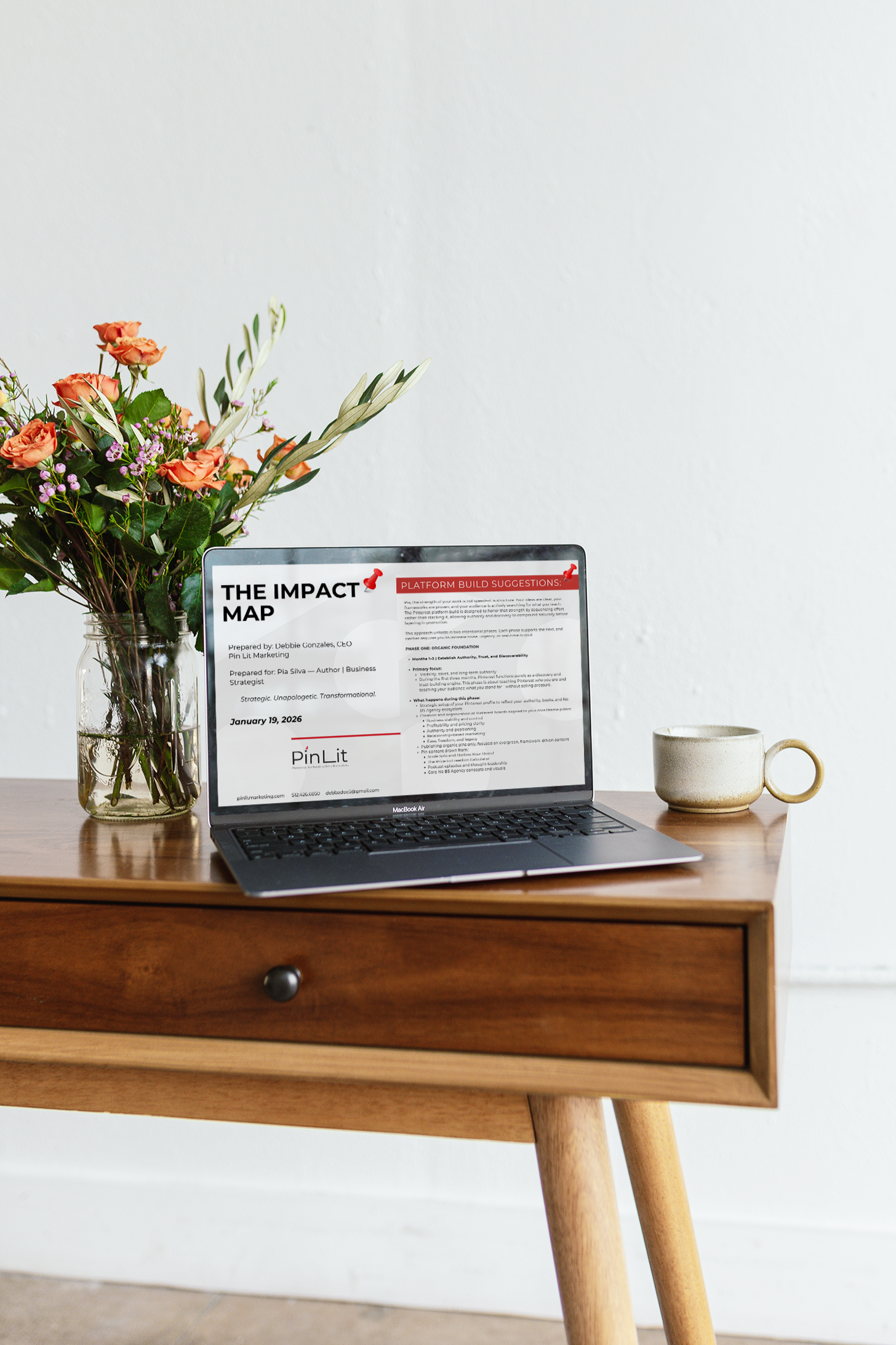 A wooden desk holding a laptop, a glass vase with pink and white flowers, and a ceramic mug against a plain white wall.