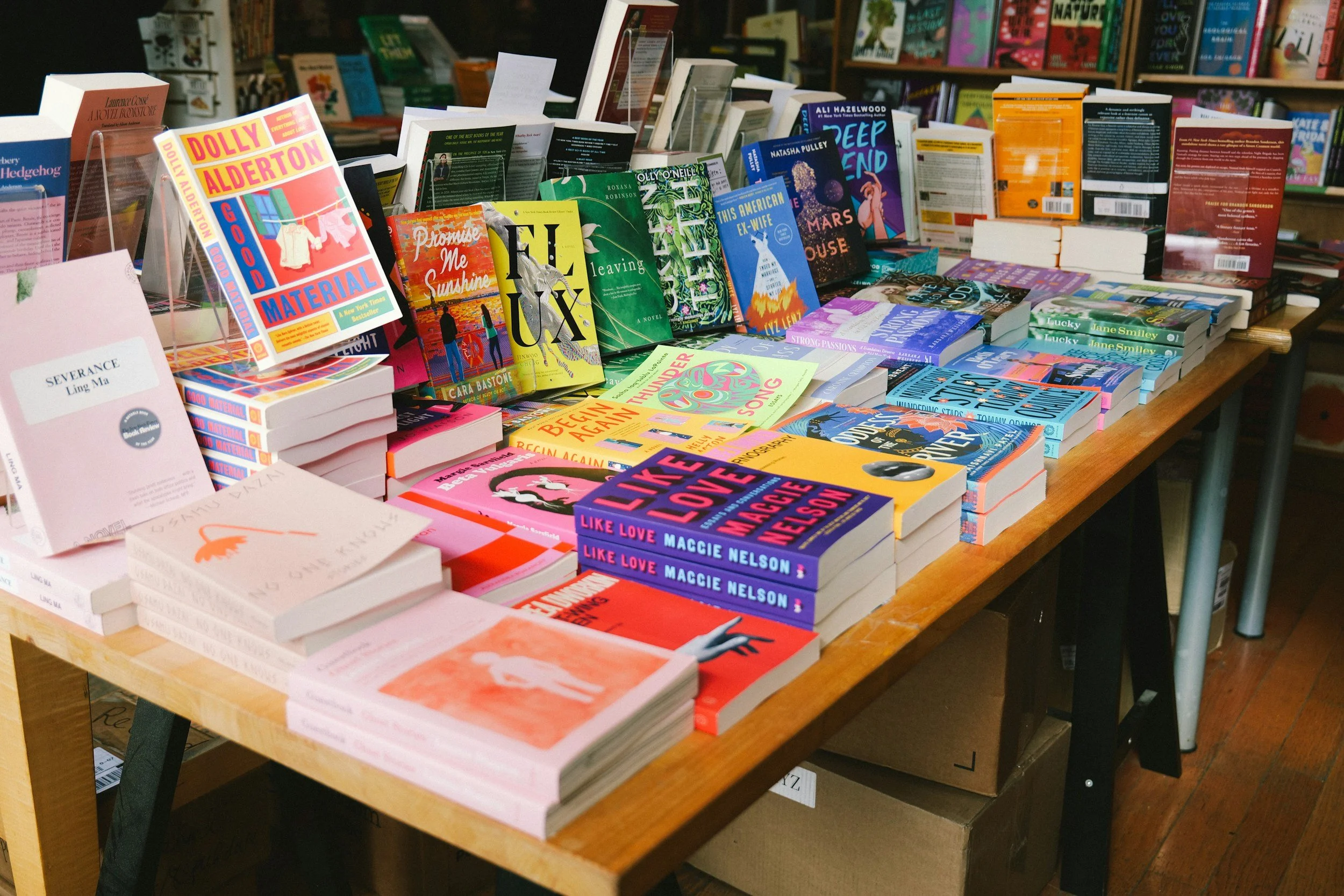 A table filled with various paperback and hardcover books at a bookstore. The books have colorful covers and are stacked in piles or arranged upright. The background shows more bookshelves with books.