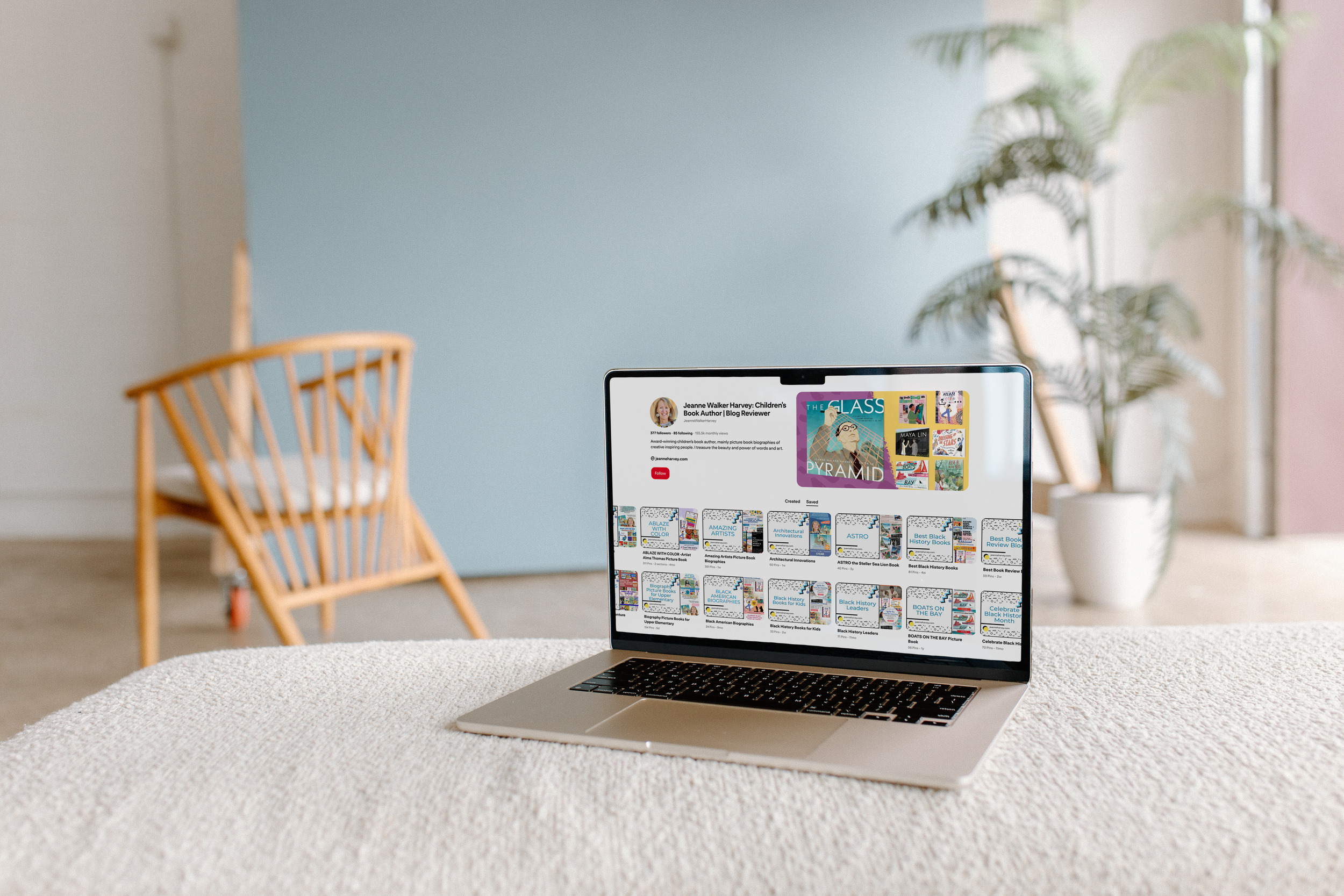A laptop on a beige textured surface displaying a Pinterest profile page, with a wooden chair and a potted plant in the background.