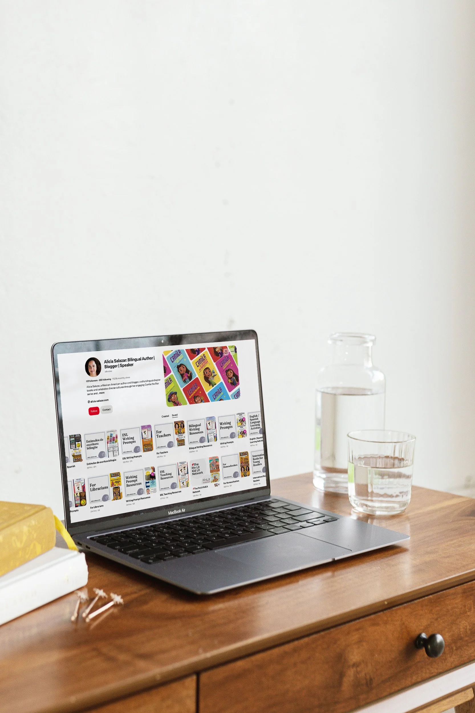A MacBook Air laptop on a wooden desk displaying a Pinterest profile of Alicia Salazar, with books and a glass pitcher of water and cup nearby.