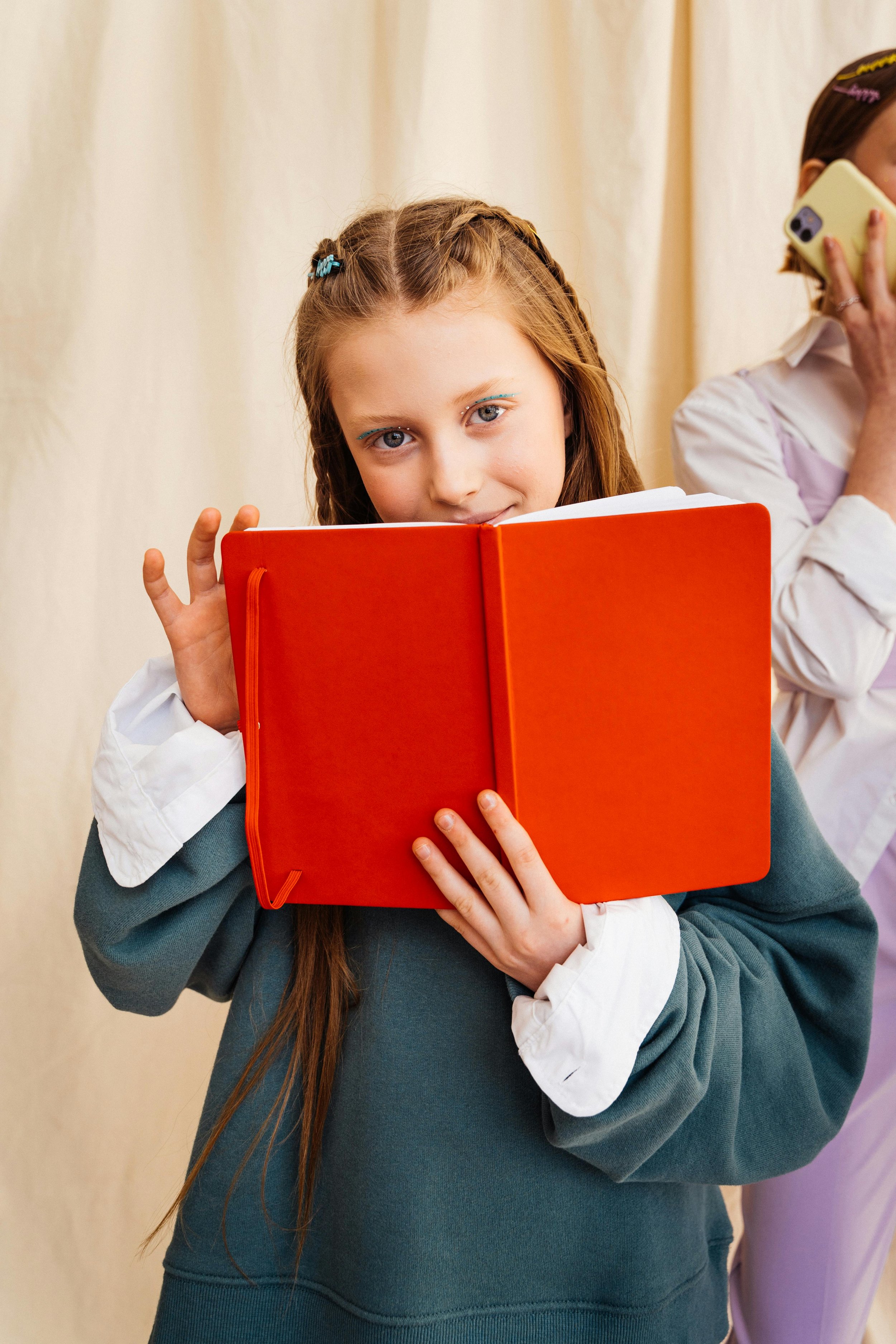 A girl with long auburn hair, wearing a blue sweatshirt with white cuffs, holds a red book close to her face with a gentle smile, standing against a cream-colored curtain background.